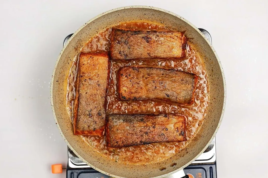 A skillet on a stove with some oil and four golden-brown salmon fillets skin-side up in it.