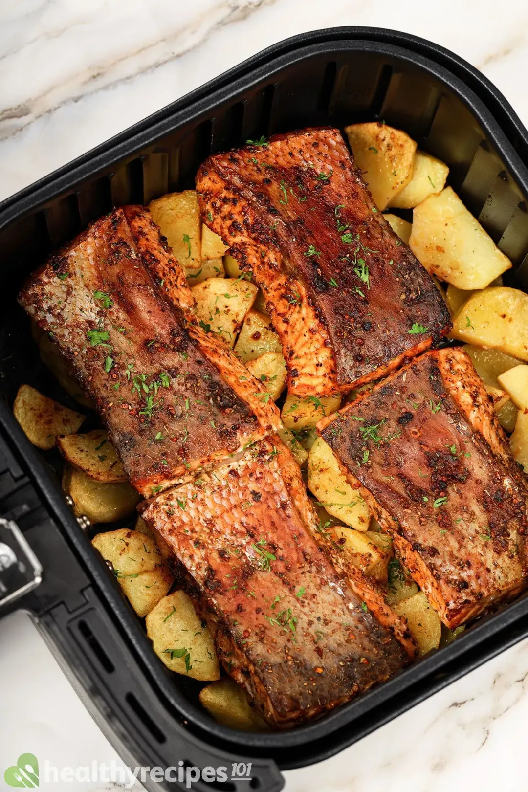 An air fryer basket containing four salmon fillets with brown skin and surrounded by large potato cubes