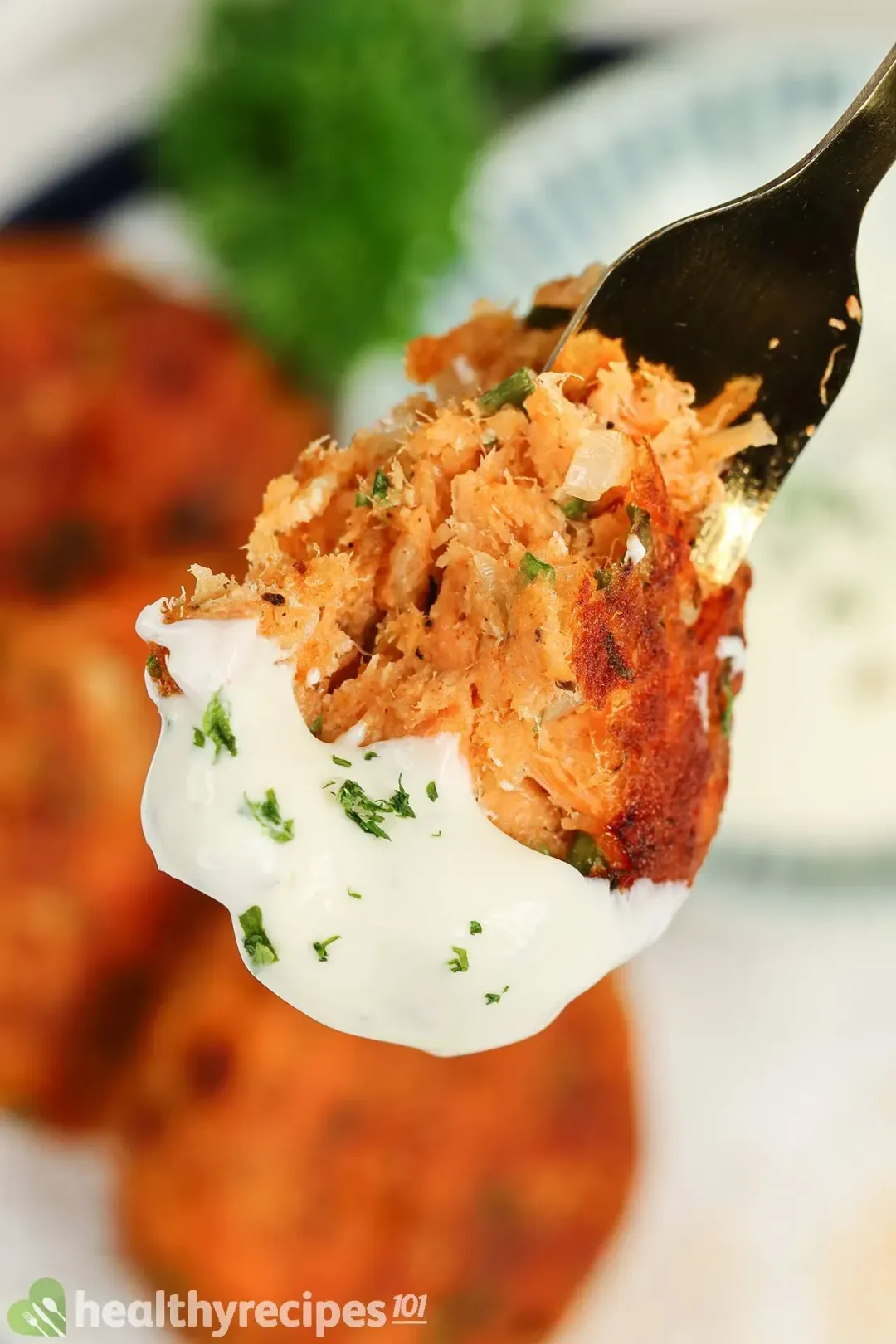 A close up picture of a piece of fried salmon patty dipped in white sauce and chopped parsley on a fork.