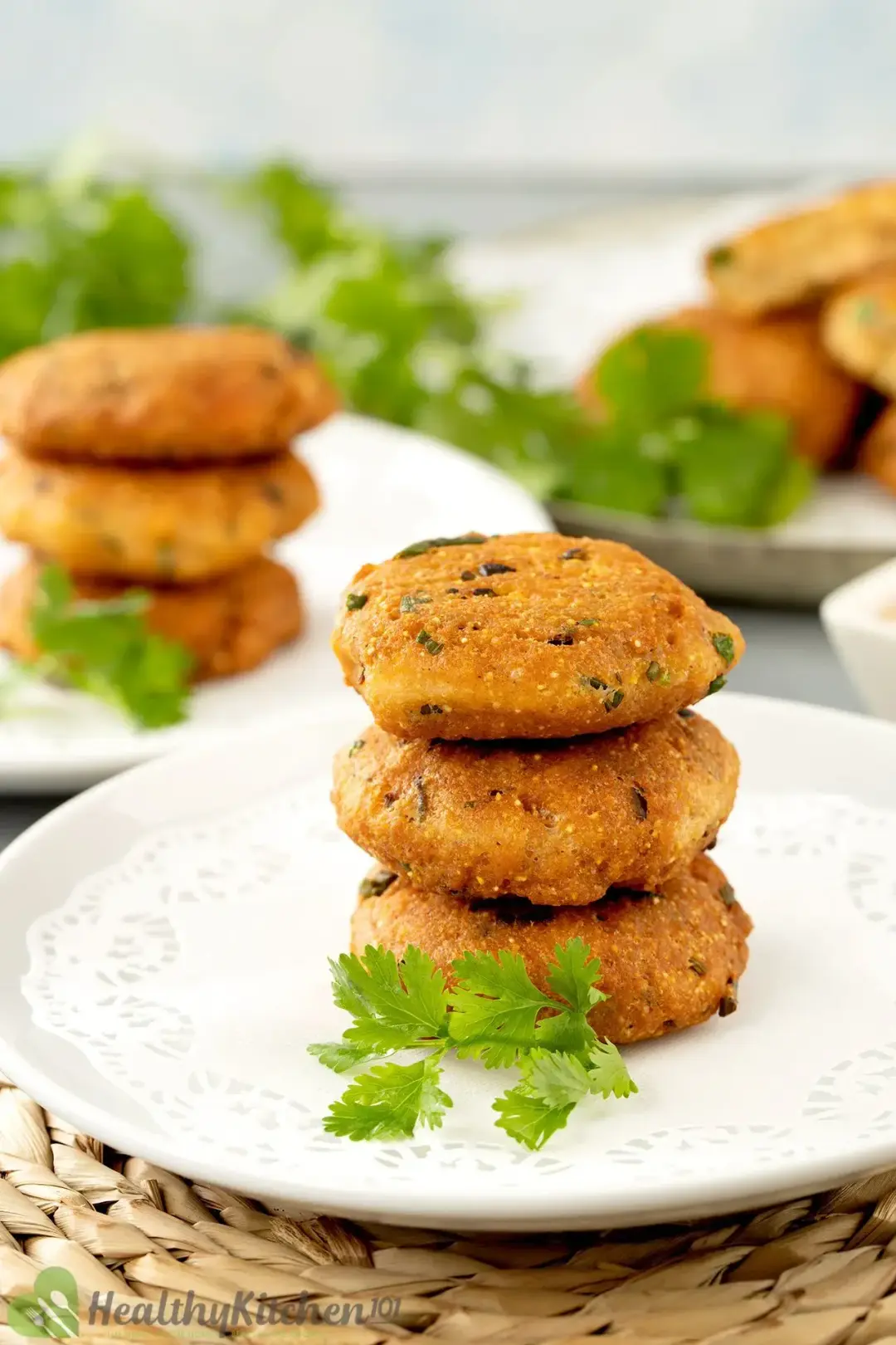 A stack of 3 golden salmon patties centering a white plate with cilantros, with greens and foods in the back