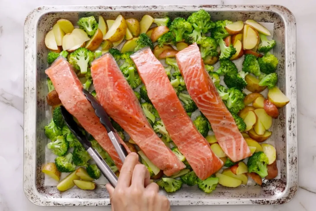 Four raw salmon filets are being placed on top of a bed of potato wedges and broccoli by a food grabber, all of which assembled on a baking sheet.