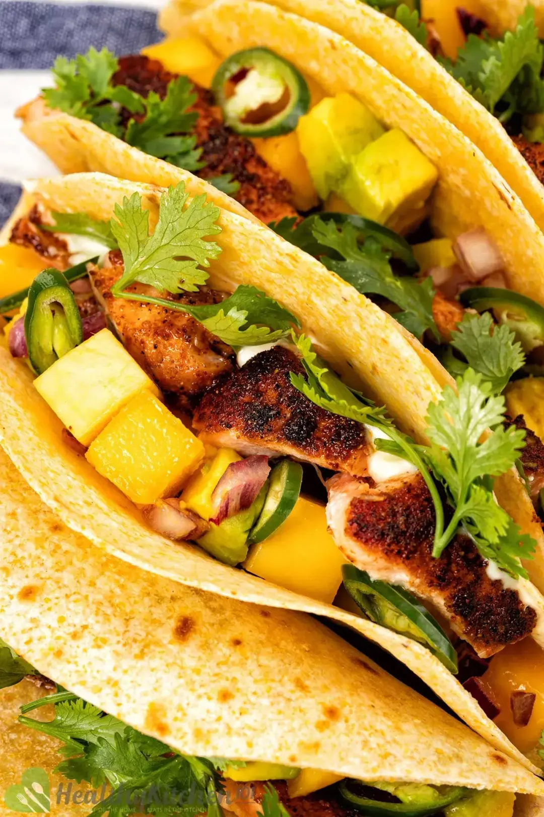 A close up picture of some blackened salmon tacos with blackened pieces of salmon, avocado cubes, and mango cubes inside crispy tortillas topped with fresh parsley.