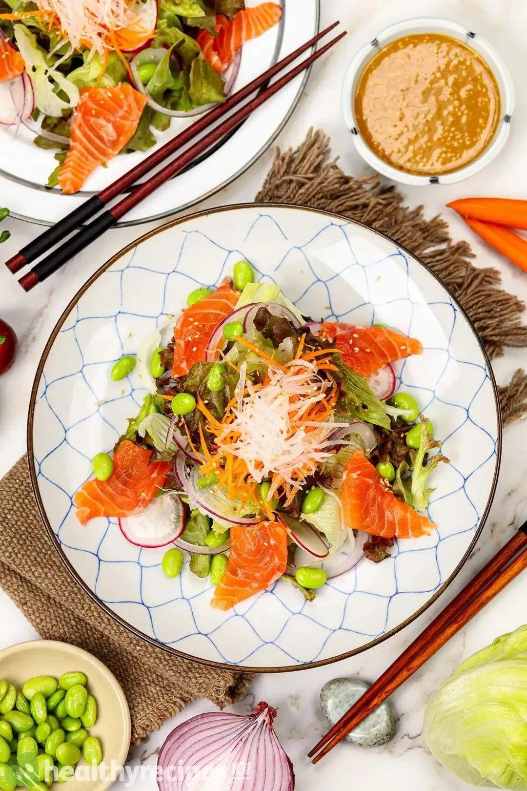 A plate of Japanese salmon salad on a tablecloth alongside another plate, a bowl of dressing, and chopsticks