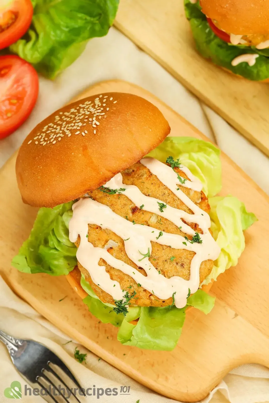 a wooden board containing a hamburger with the top bun fallen on the side, revealing a pattie covered in a sauce and chopped herbs