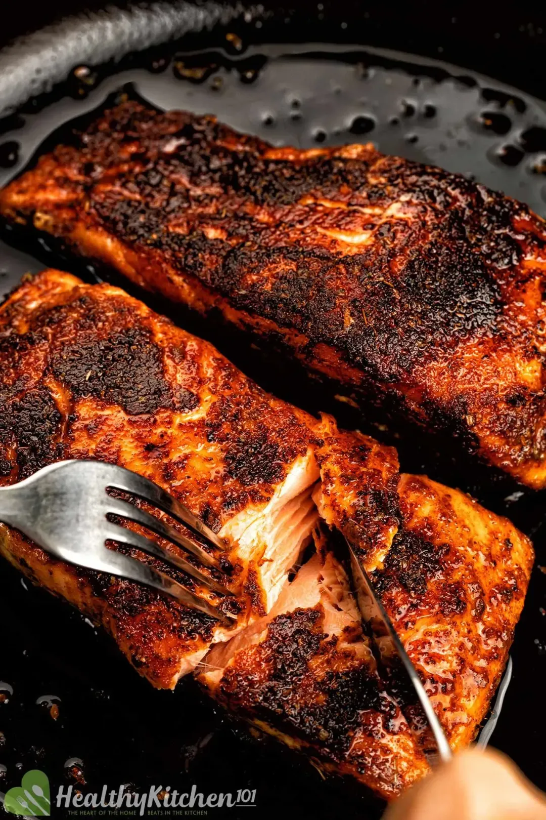 A close up picture of a fork and a knife cutting one of the two blackened salmon fillets in a black skillet.