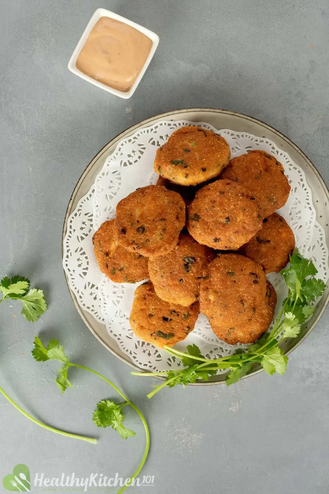 A white plate full of salmon patties with fresh cilantro sprigs and a square pink mayo dipping sauce