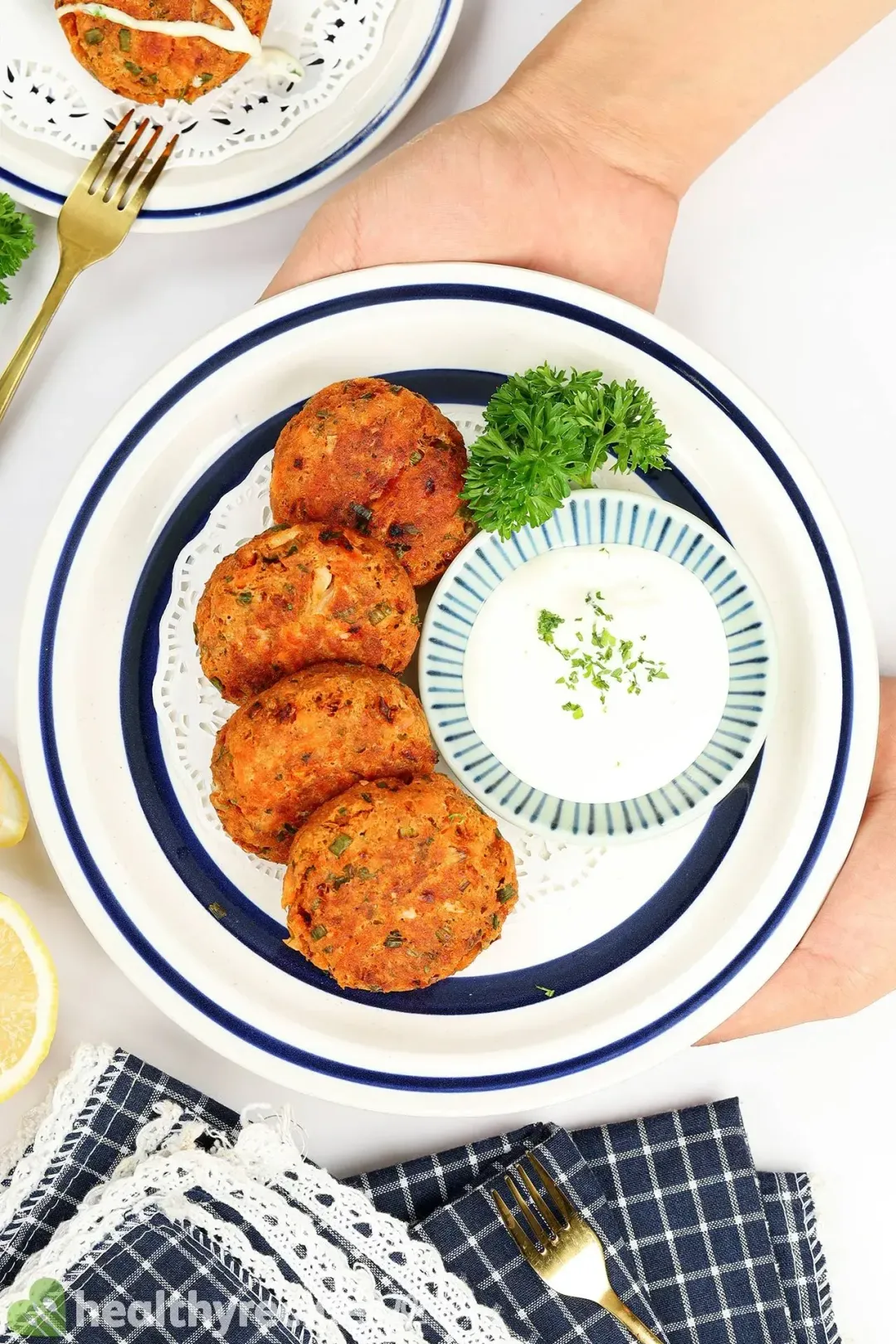 two hand holding a plate of four fried salmon patties with a small bowl of white sauce and some fresh parsley