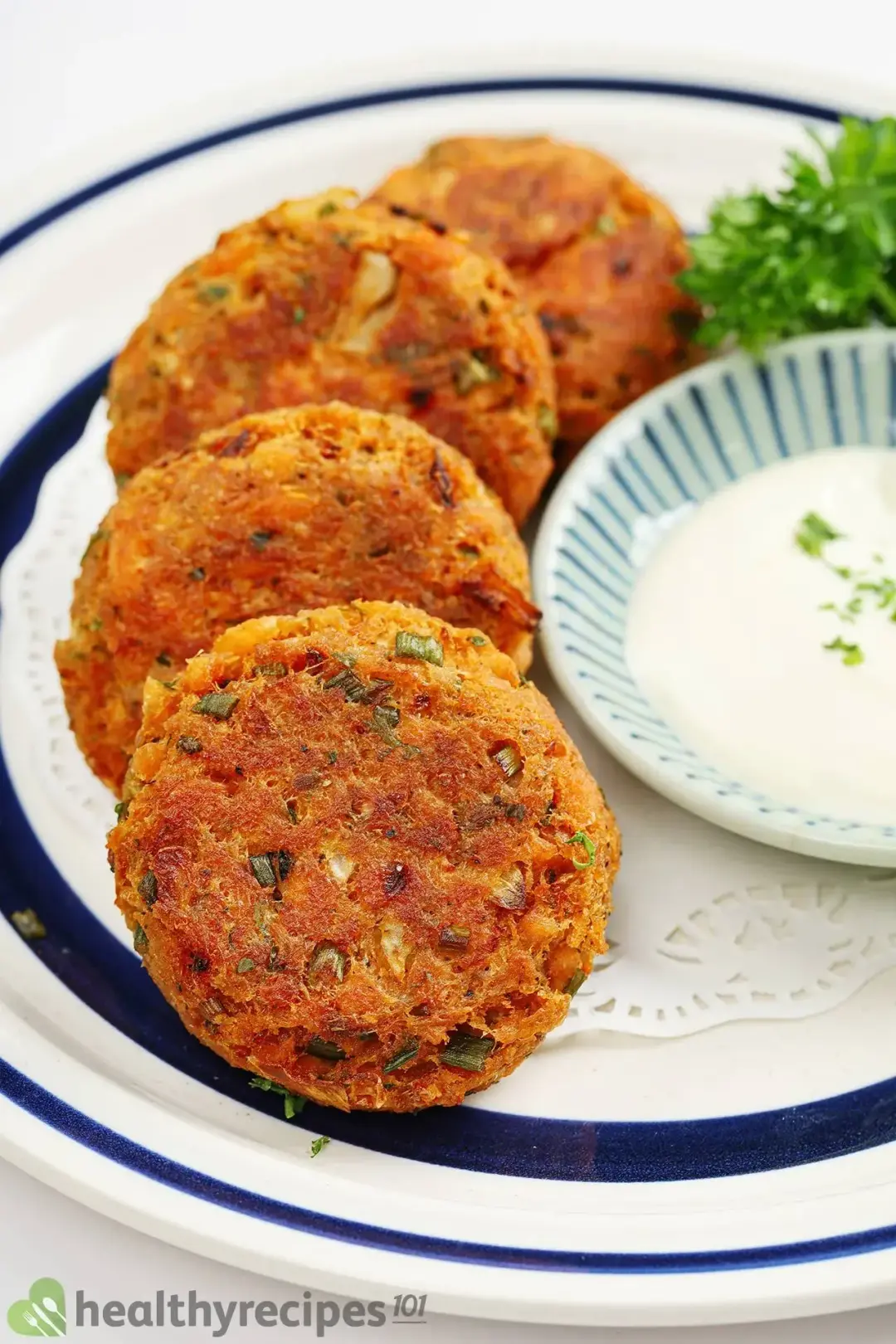 A close up picture of four fried salmon patties on a plate with a small bowl of white sauce and some fresh parsley.
