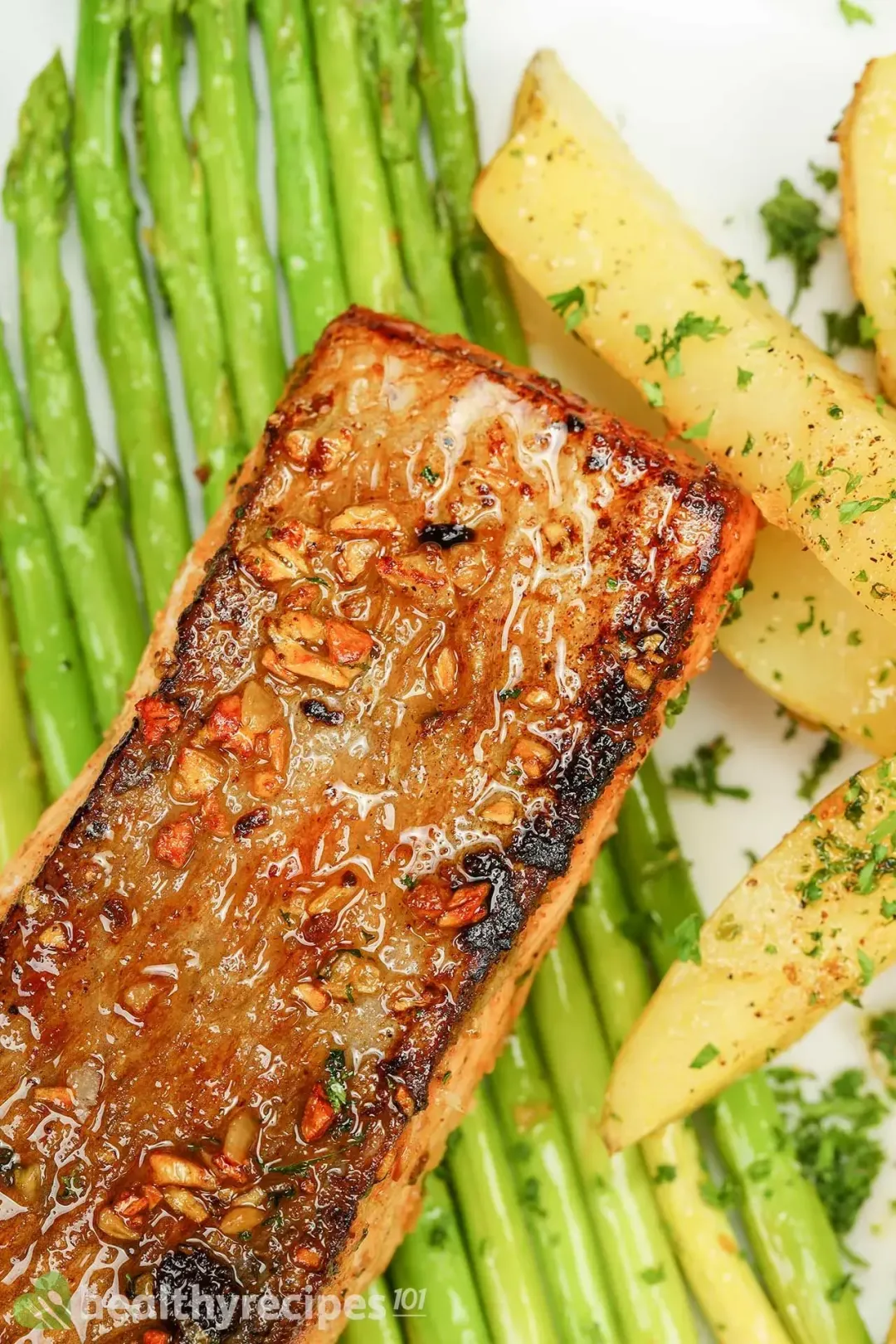 a plate containing a pan-seared salmon fillet, potato wedges covered in parsley, asparagus stalks