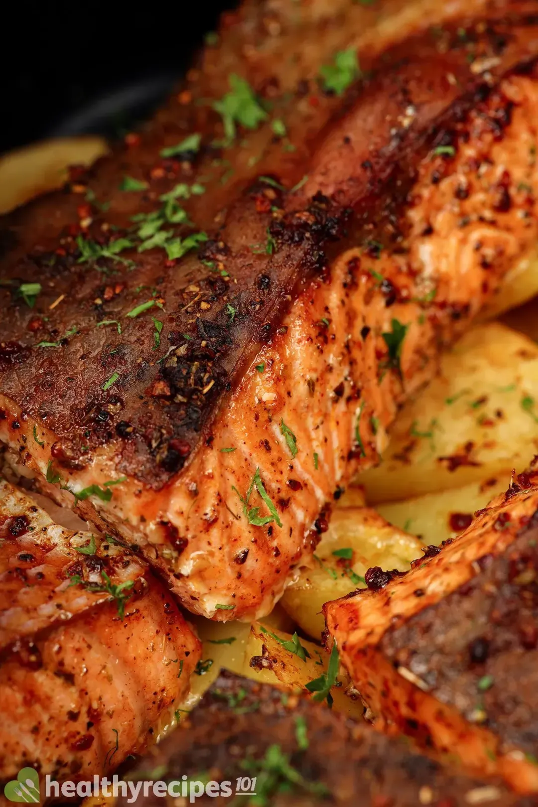 A close-up shot of a well-cooked salmon fillet with bright orange flesh and charred brown skin covered in chopped herbs and laid over potato cubes