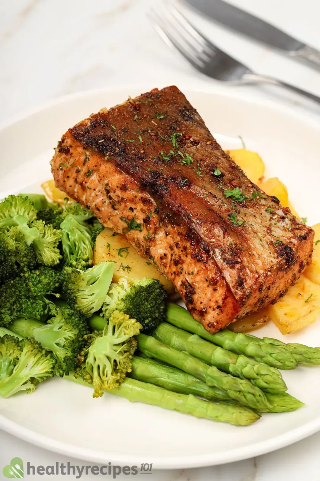 A plate of air fryer salmon, potato, broccoli florets, and asparagus laid near a silver spoon and fork