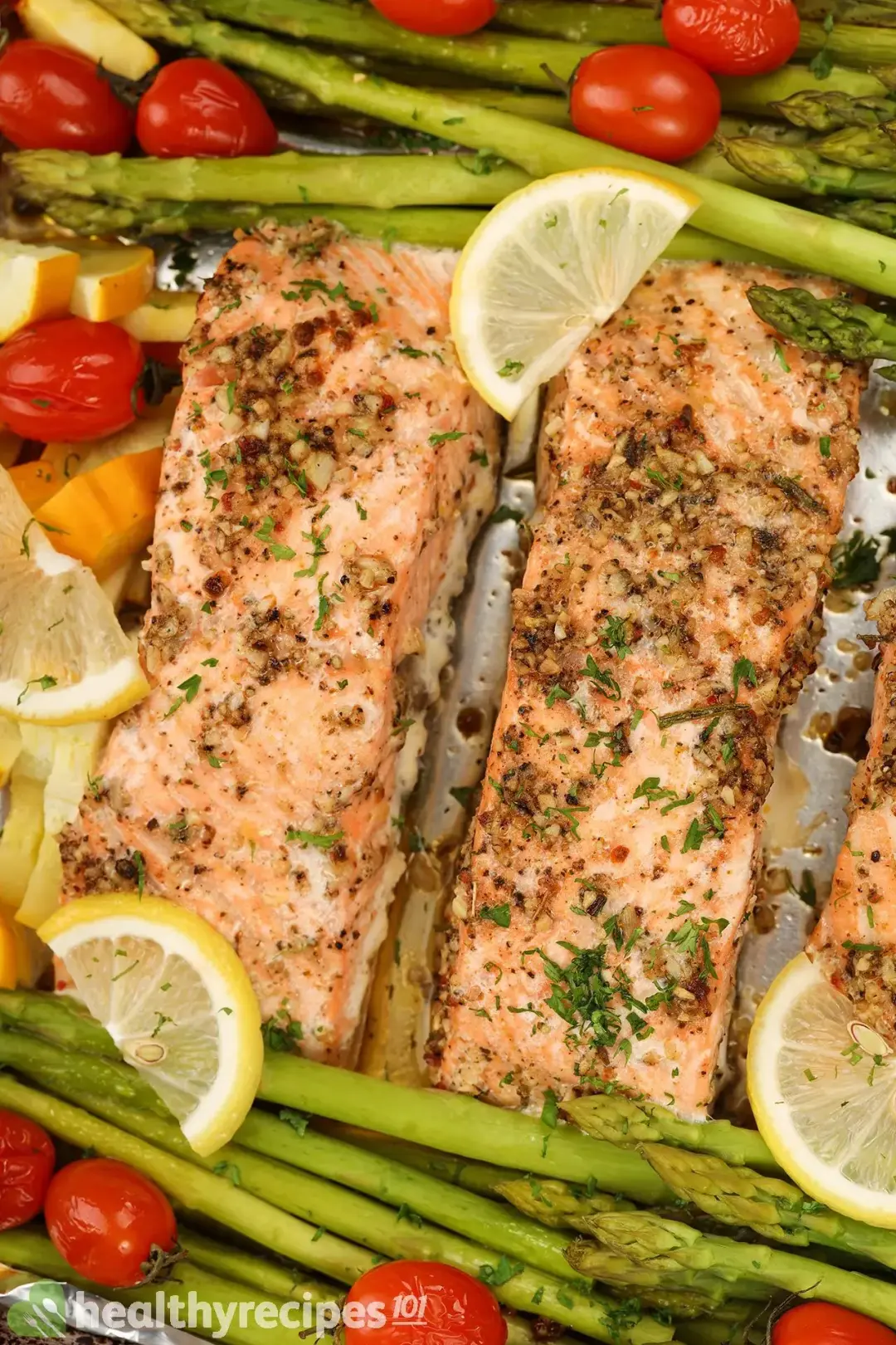 A close-up picture of baked salmon fillets, cherry tomatoes, asparagus, zucchini, and lemon slices on a baking tray taken from above