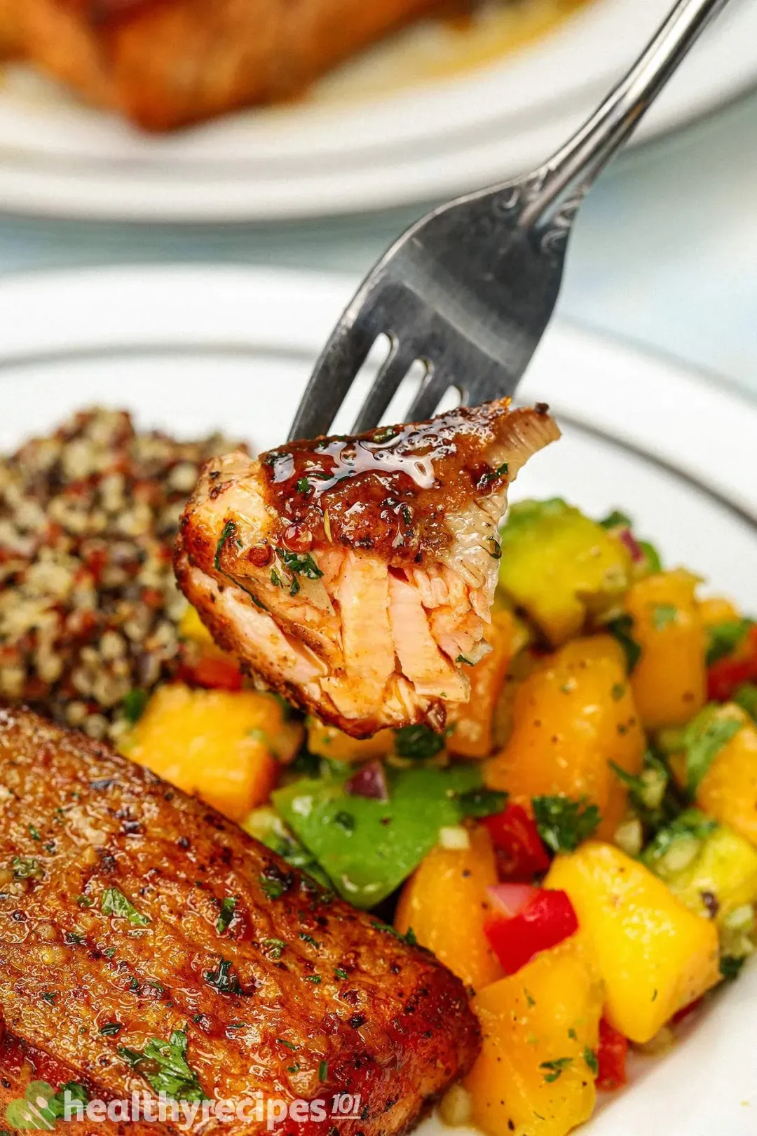 A close up photo of a piece of salmon in the background of a plate of pan-seared salmon fillet, quinoa, and mango-avocado salsa.