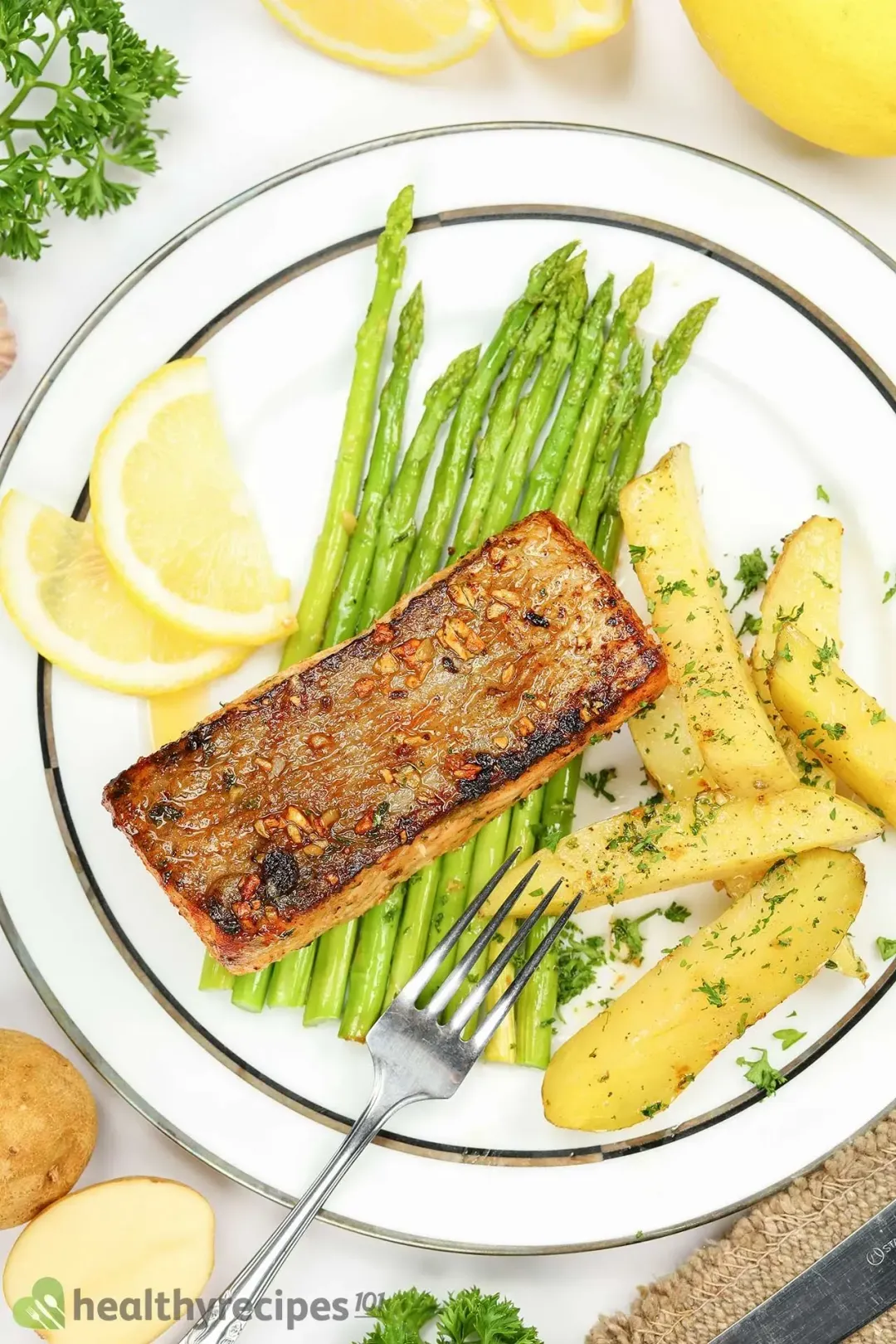 A fork laid on a plate containing a pan-seared salmon fillet, potato wedges covered in parsley, asparagus stalks, and two lemon slices