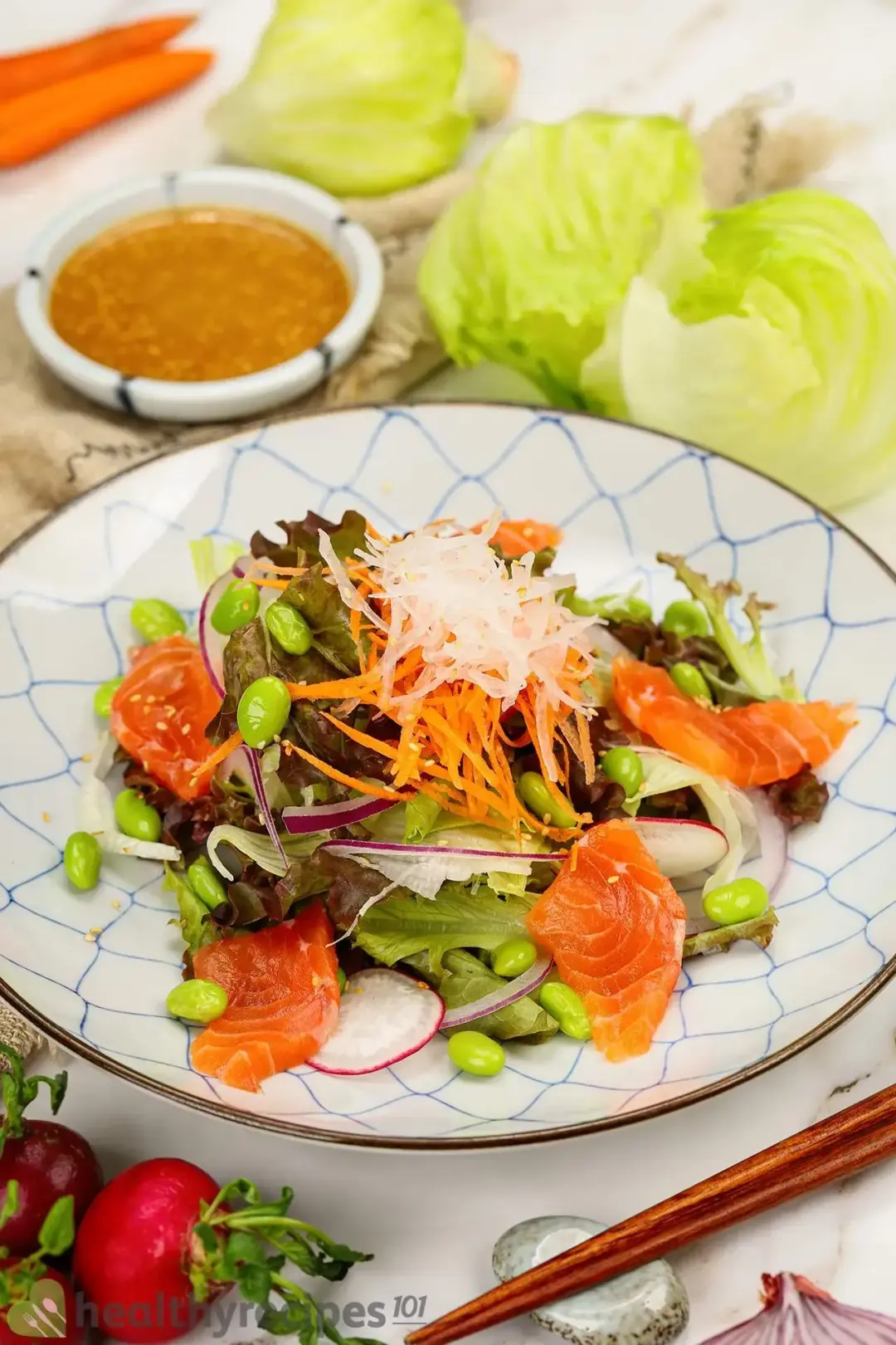 A close-up shot of a plate of Japanese salmon salad next to a bowl of dressing, a pair of chopsticks, radish, carrots, and cabbage
