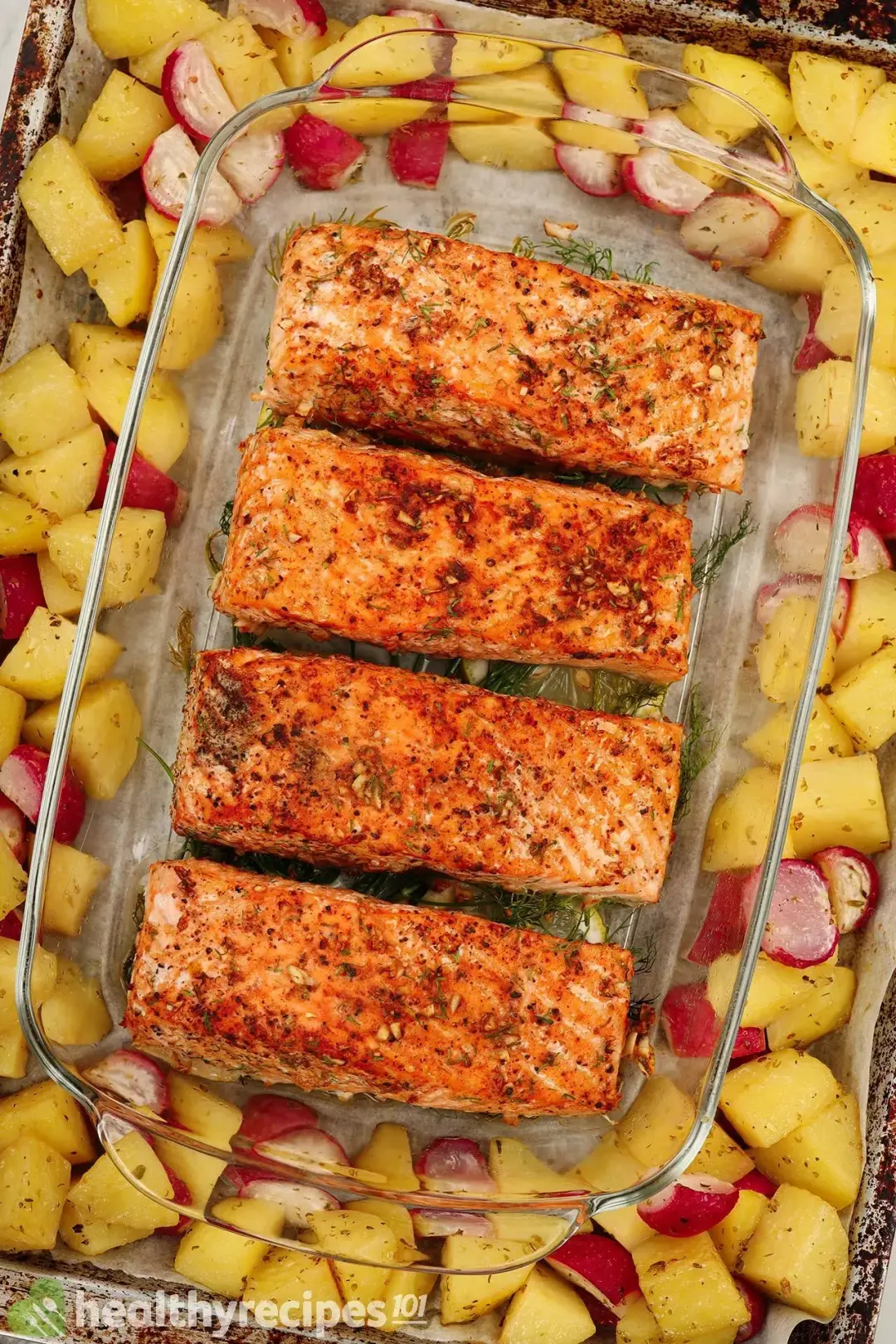 A high-angle shot of a glass tray containing four cooked salmon fillets surrounded by potato cubes and radish cubes. Everything is laid on a baking sheet lined with parchment paper