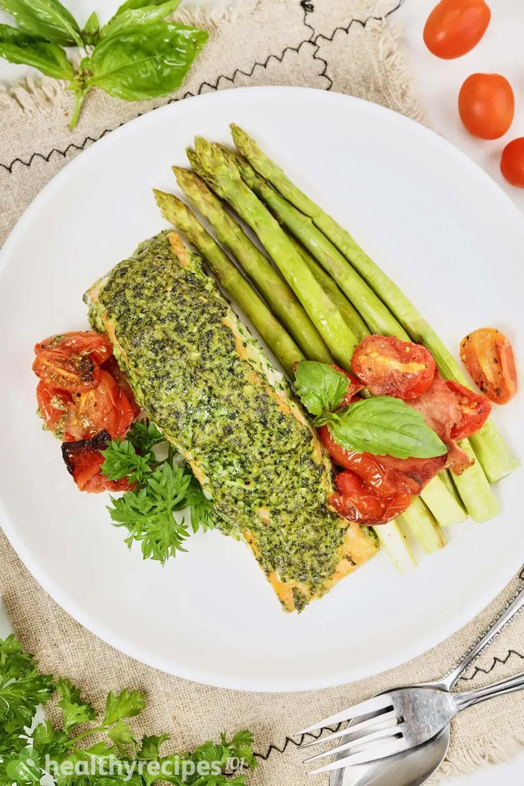 A plate of salmon pesto on a tablecloth surrounded by some fresh basil leaves, cherry tomatoes, parsley
