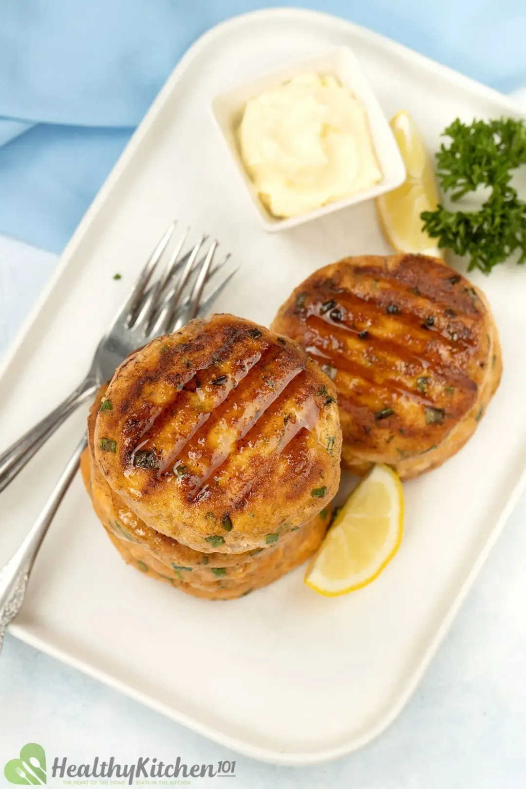 A picture taken from above of a rectangular plate of salmon croquettes with a small square bowl of white sauce, together with some forks, lemon slices, and parsley for decoration.