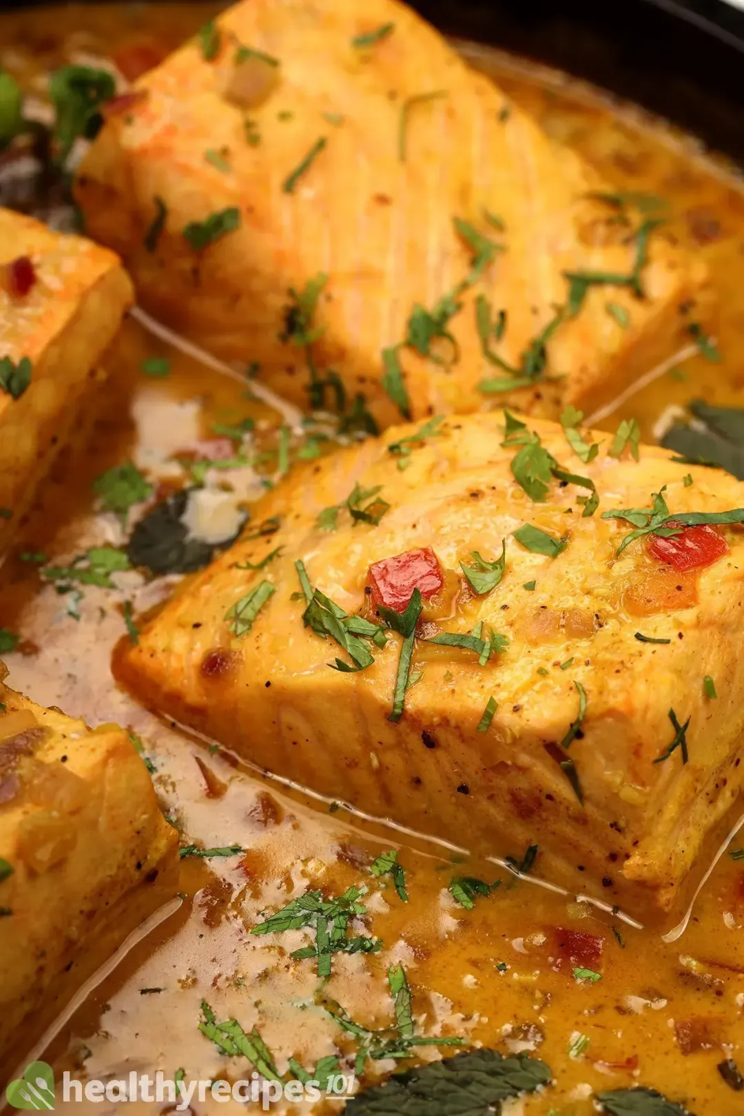 A close-up shot of a cooked salmon fillet covered in yellow curry, chopped herbs, and diced tomatoes