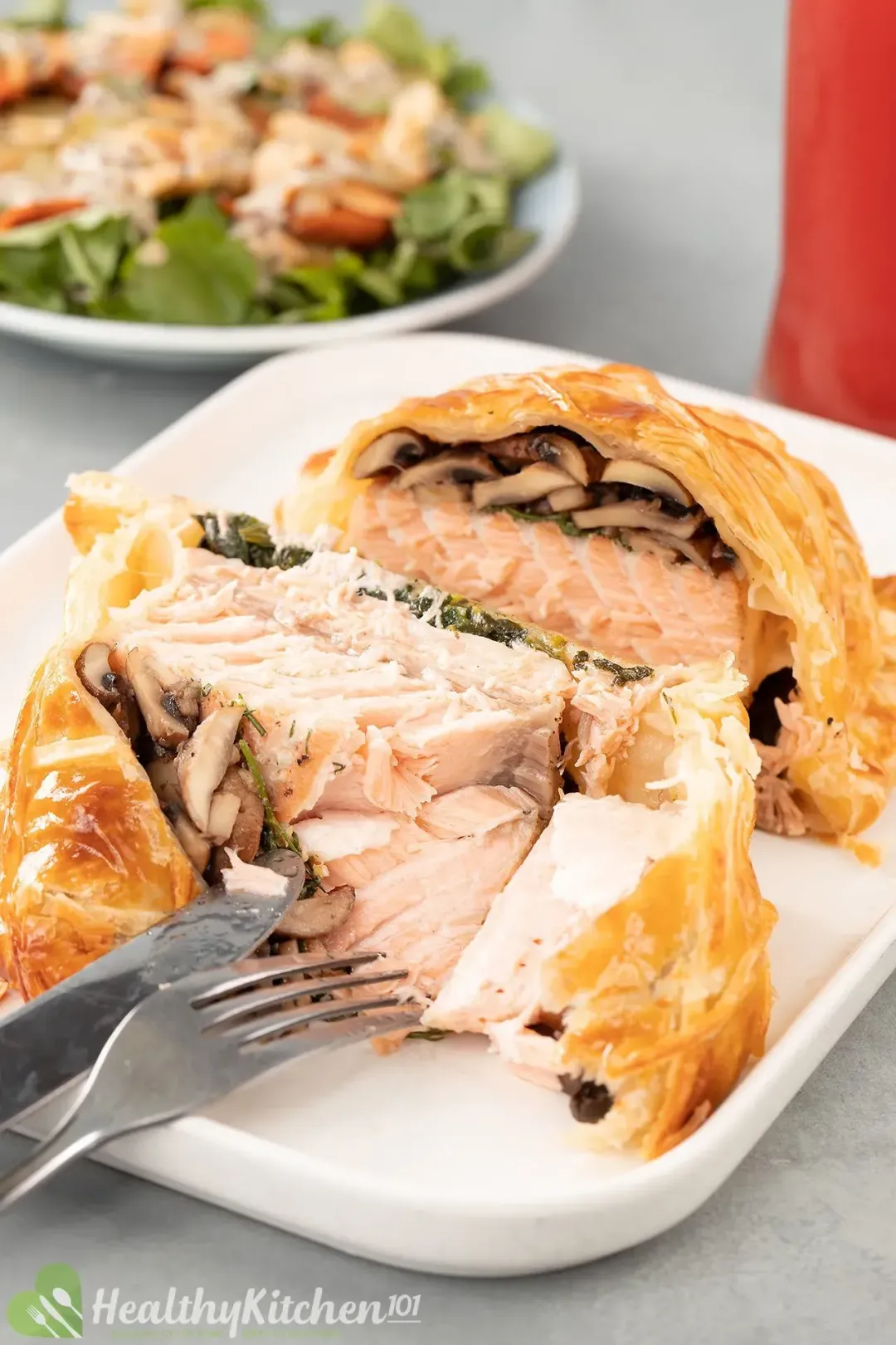 A plate containing salmon en croute sliced open revealing cooked salmon layered with cooked mushroom covered in golden cooked puff pastry