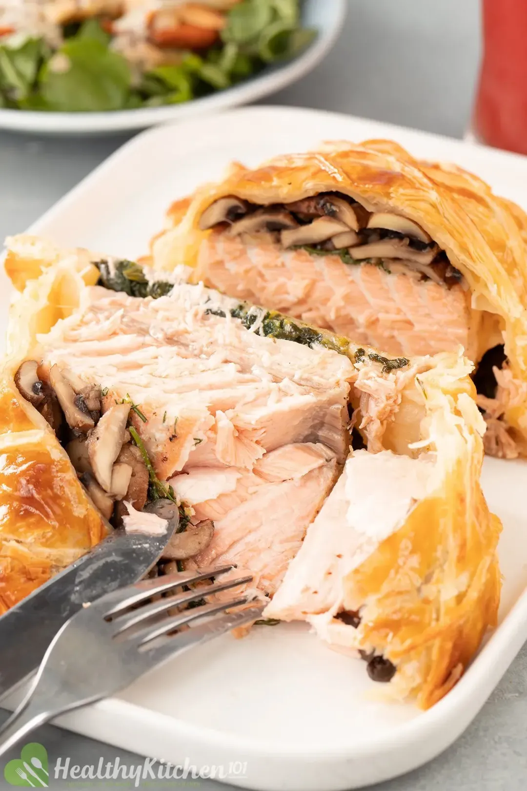 A plate containing salmon en croute sliced open revealing cooked salmon layered with cooked mushroom covered in golden cooked puff pastry