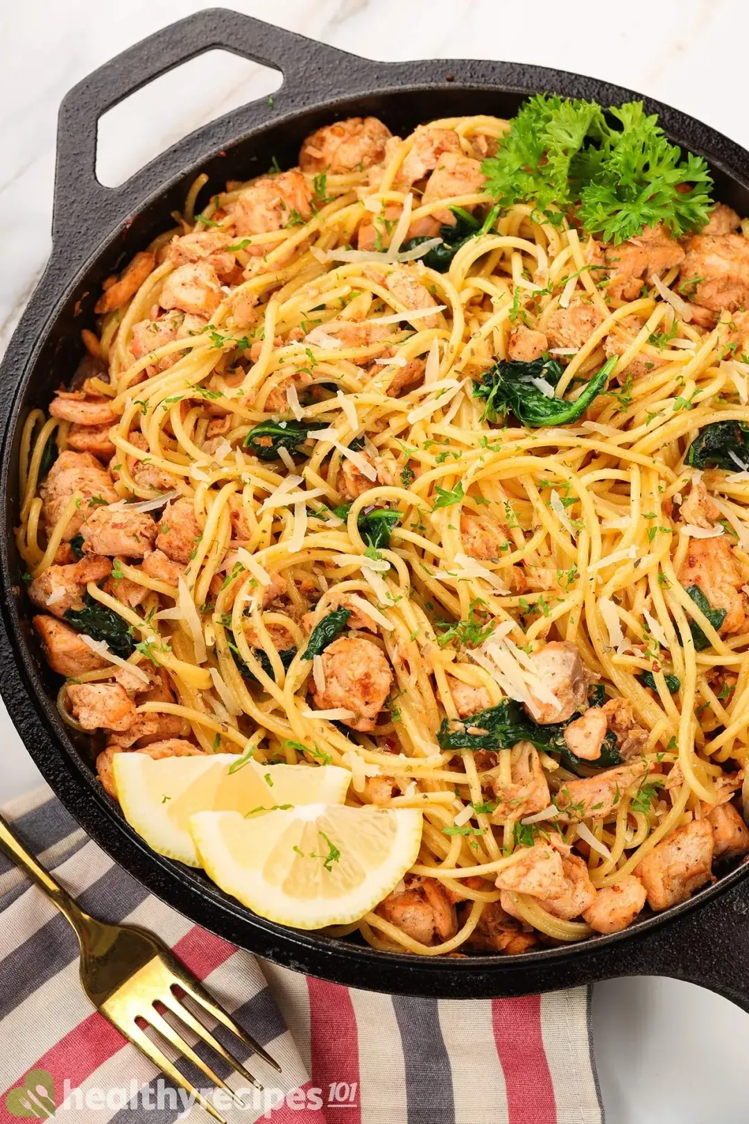 A cast iron skillet filled with cooked spaghetti, salmon, and spinach laid near a striped tablecloth, two lemon wedges, and fresh parsley