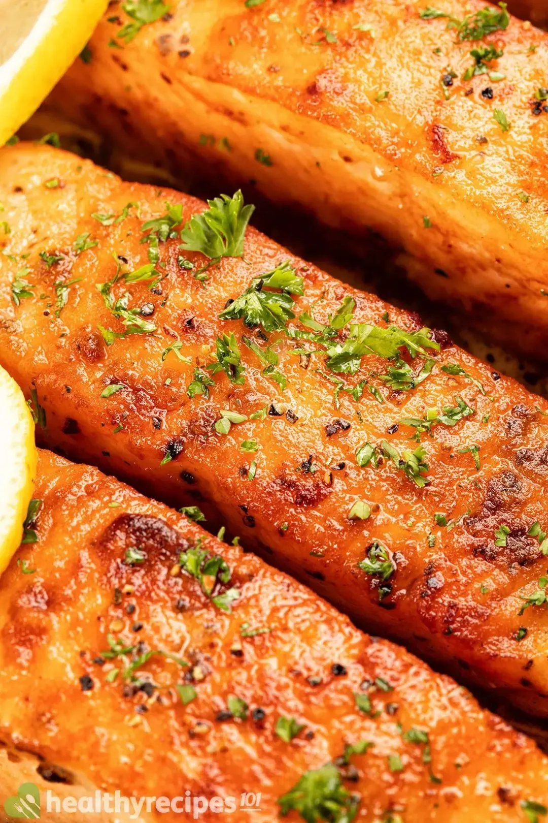 A super close-up view of the surface of some nicely browned salmon filets, sprinkled with chopped parsley and lemon slices.