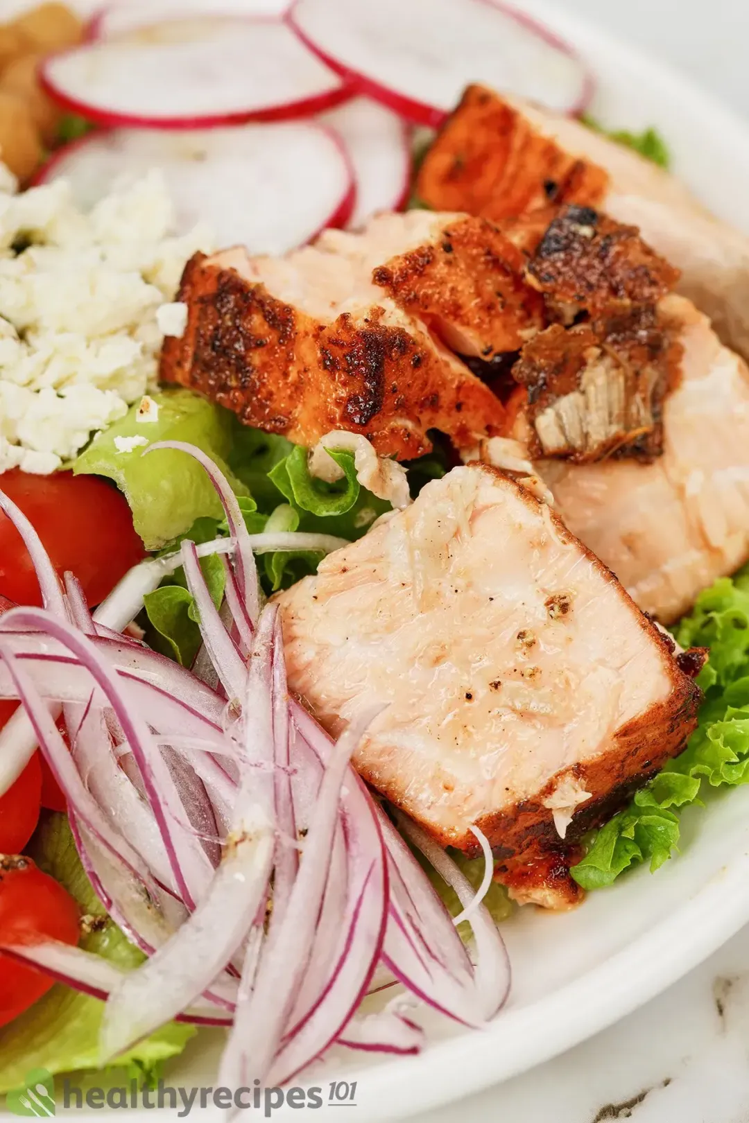 A close-up shot of pan-seared salmon fillet cubes, red onion straws, and cherry tomatoes laid on lettuce leaves