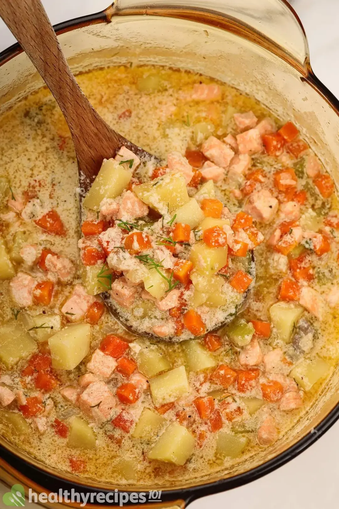 A high-angle shot of a saucepan filled with salmon soup, featuring diced vegetables and cubed salmon. There’s a wooden ladle picking up some of the food