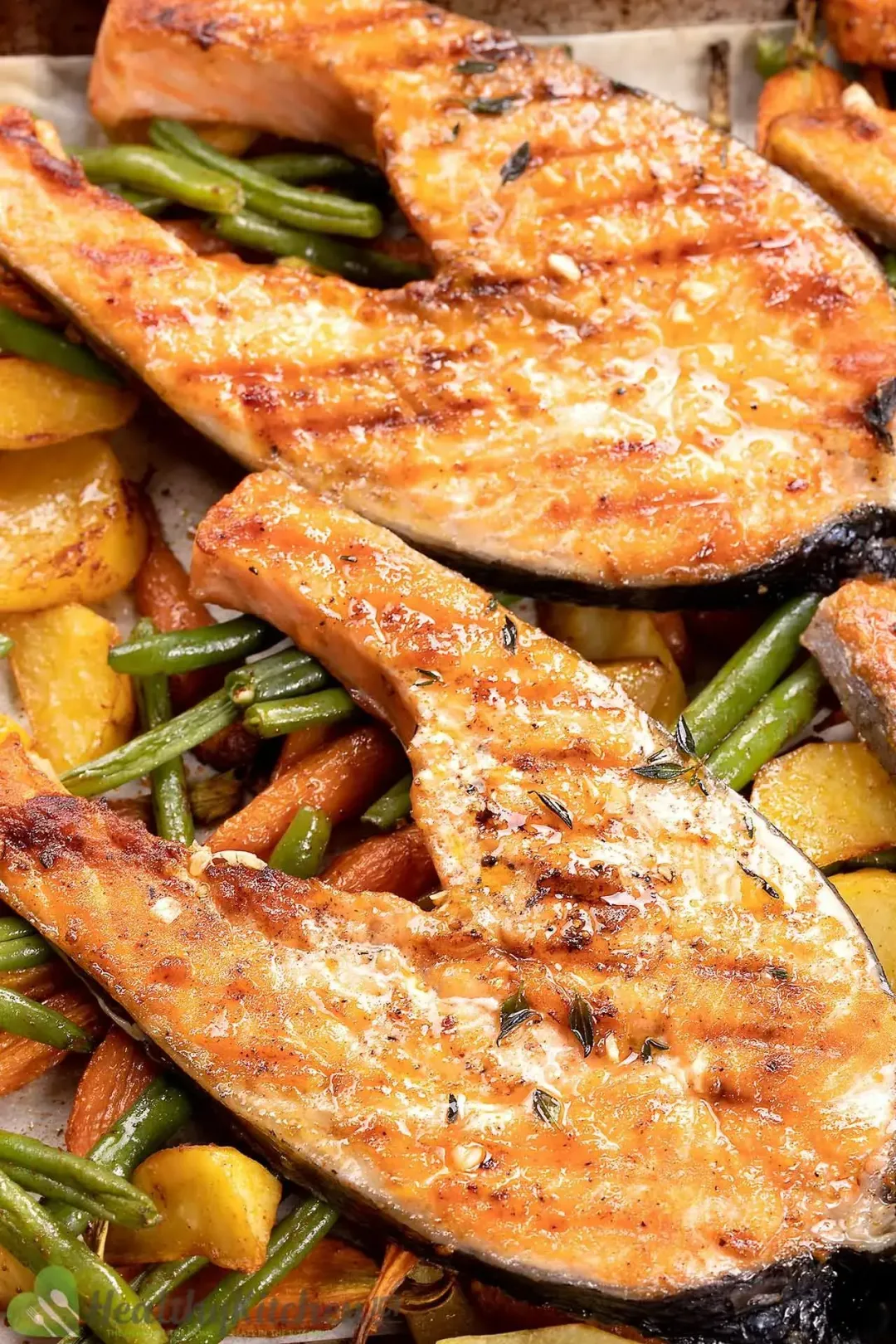 Two grilled salmon steaks atop a baking tray filled with string beans, potato wedges, and baby carrots