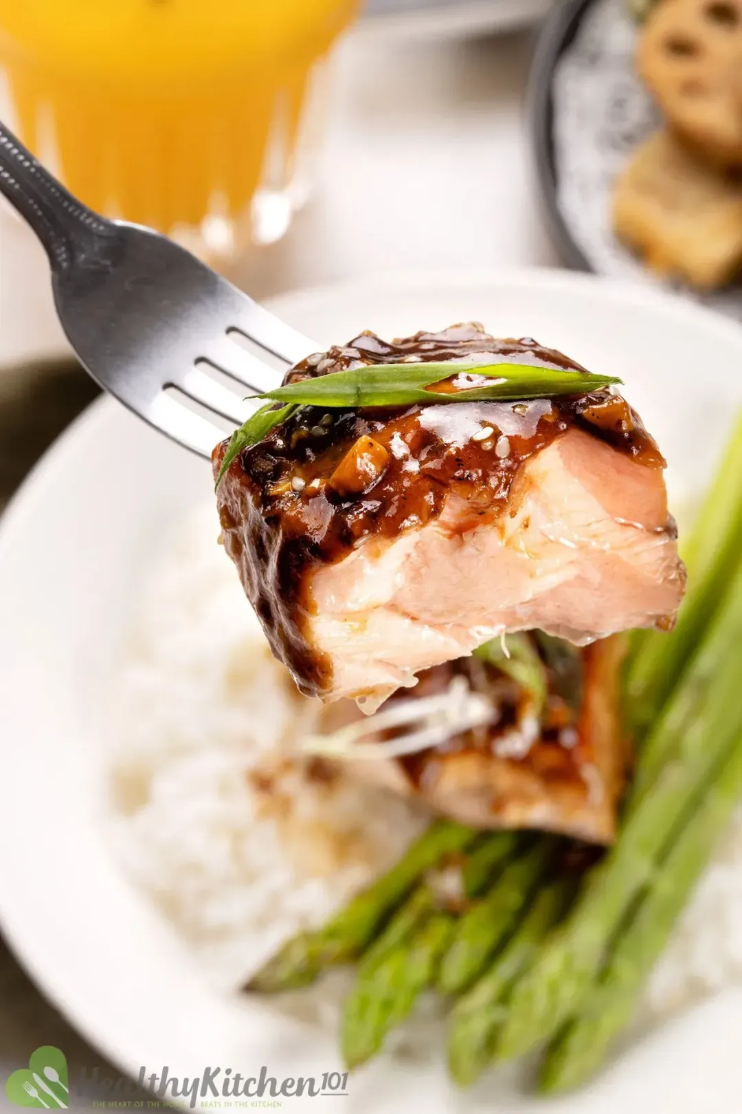 A mouth-sized piece of baked teriyaki salmon pierced by a fork with a chop of scallion placed on top; at the back is a serving plate containing a whole salmon filet, cooked white rice, and asparagus.
