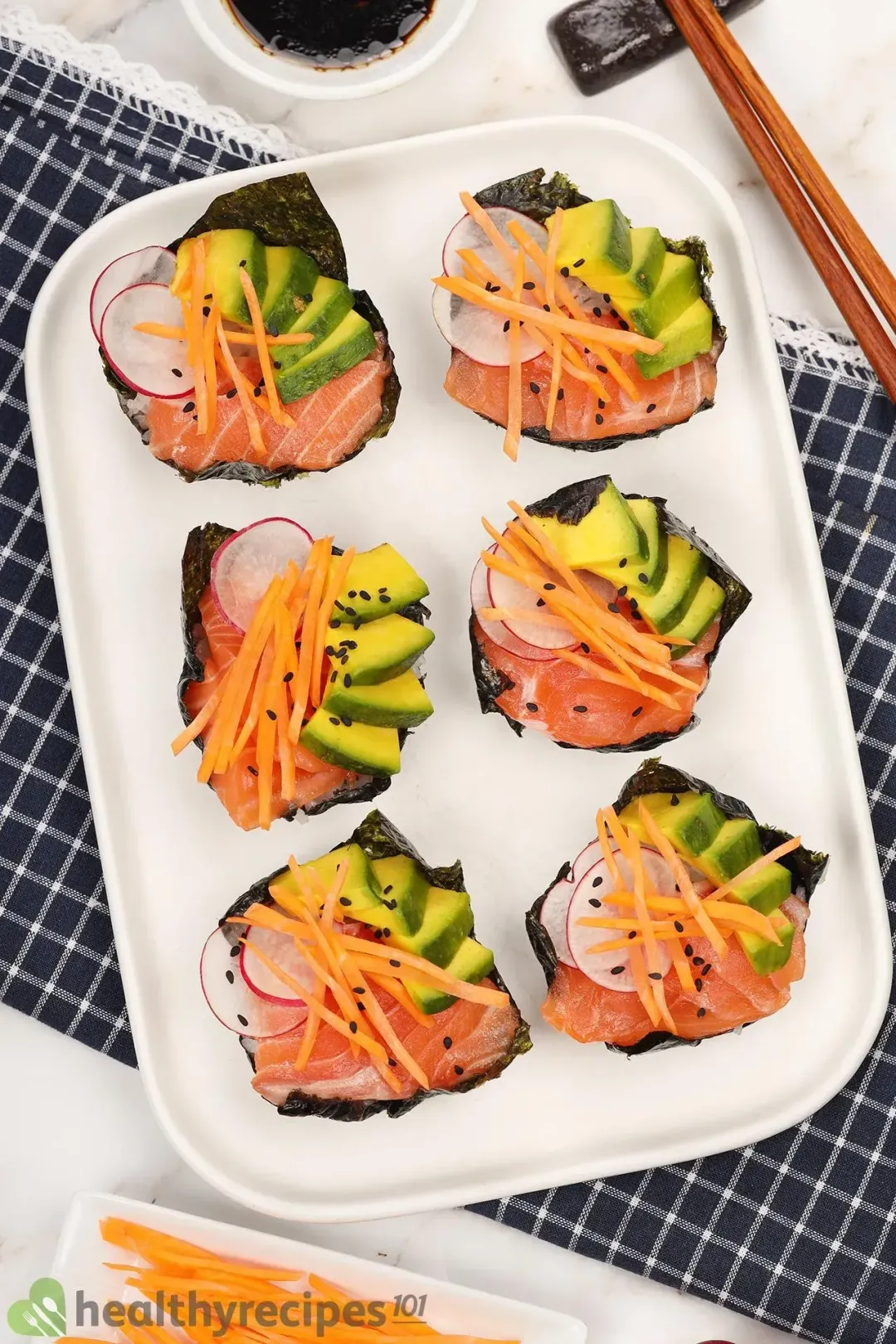 Six sushi cupcakes on a white plate, with a blue checkered cloth beneath and next to a pair of chopsticks
