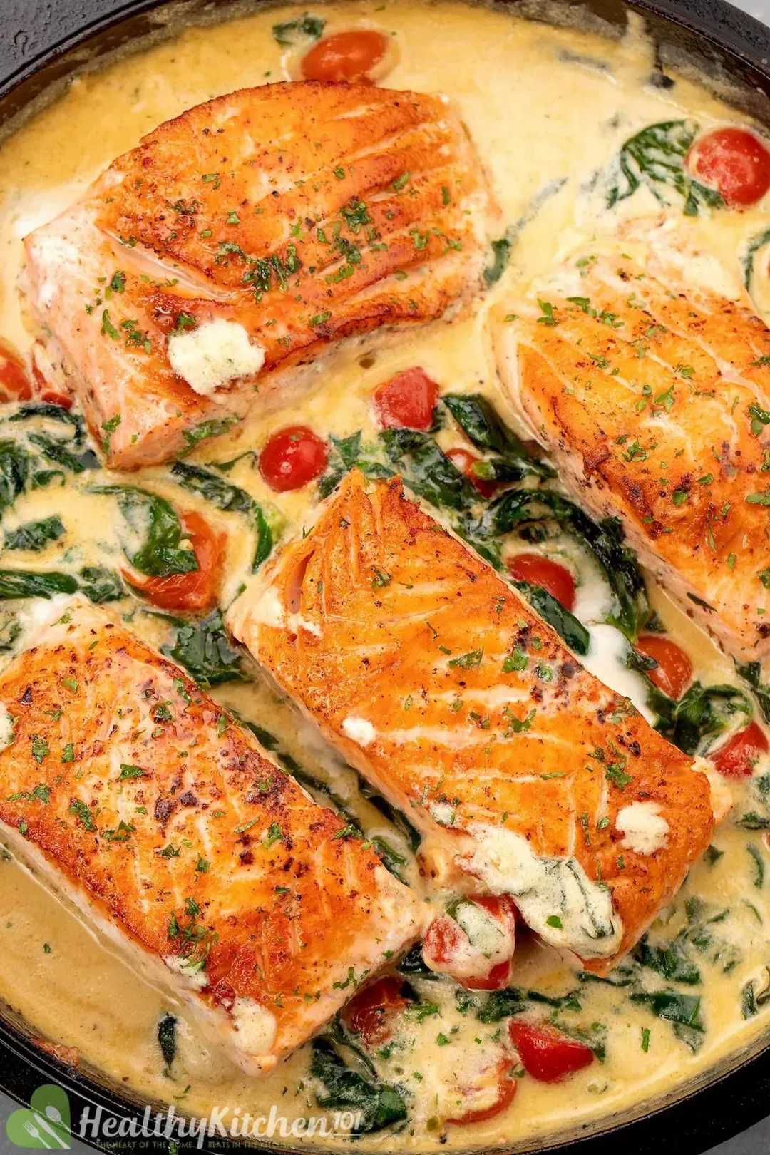 A cast iron skillet of four golden salmon filet in a cream sauce with cooked spinach and halved cherry tomatoes