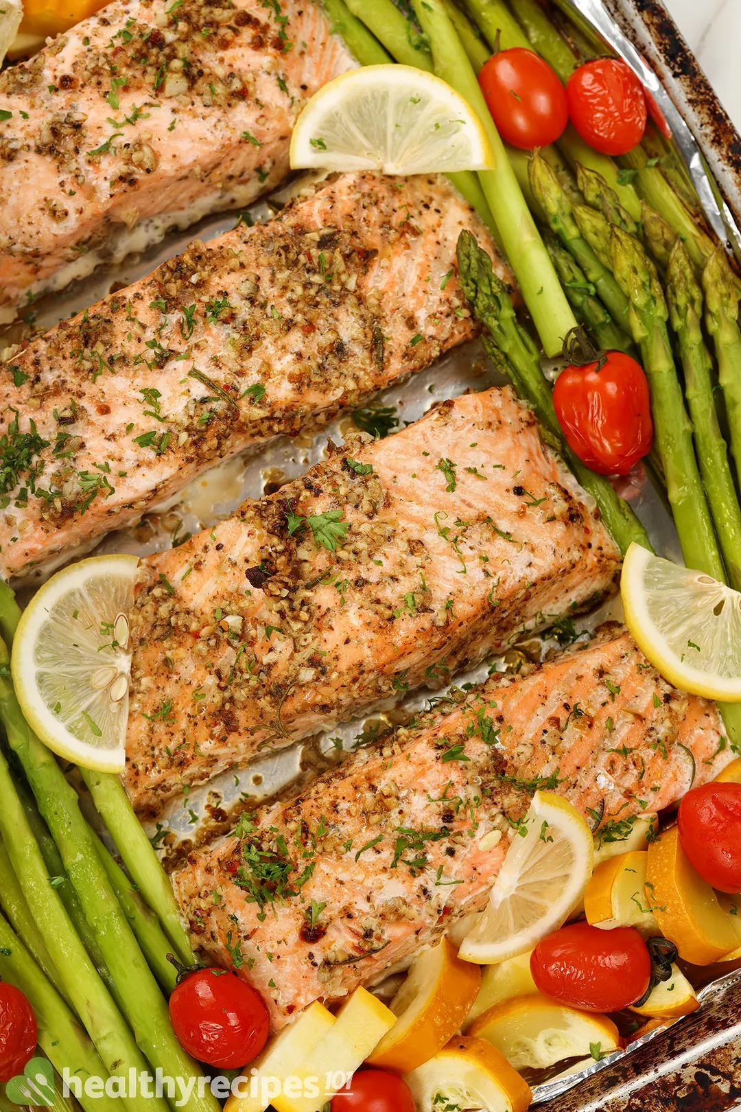 four baked salmon fillets on a tray with asparagus, tomatoes, lemon slices