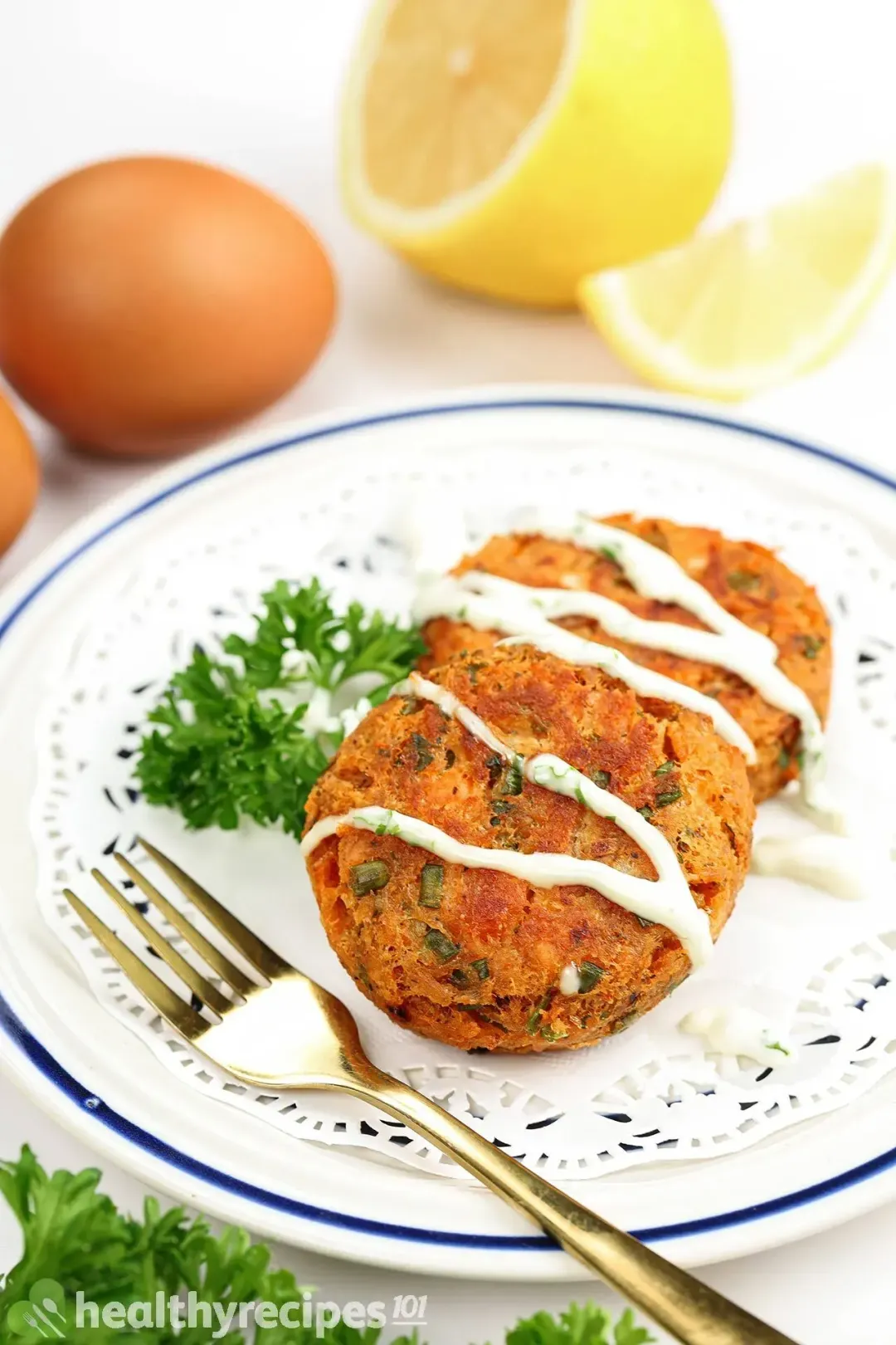A picture of a plate with two fried salmon patties drizzled with yogurt sauce and garnished with fresh parsley. There are some eggs and lemons blurred in the background.