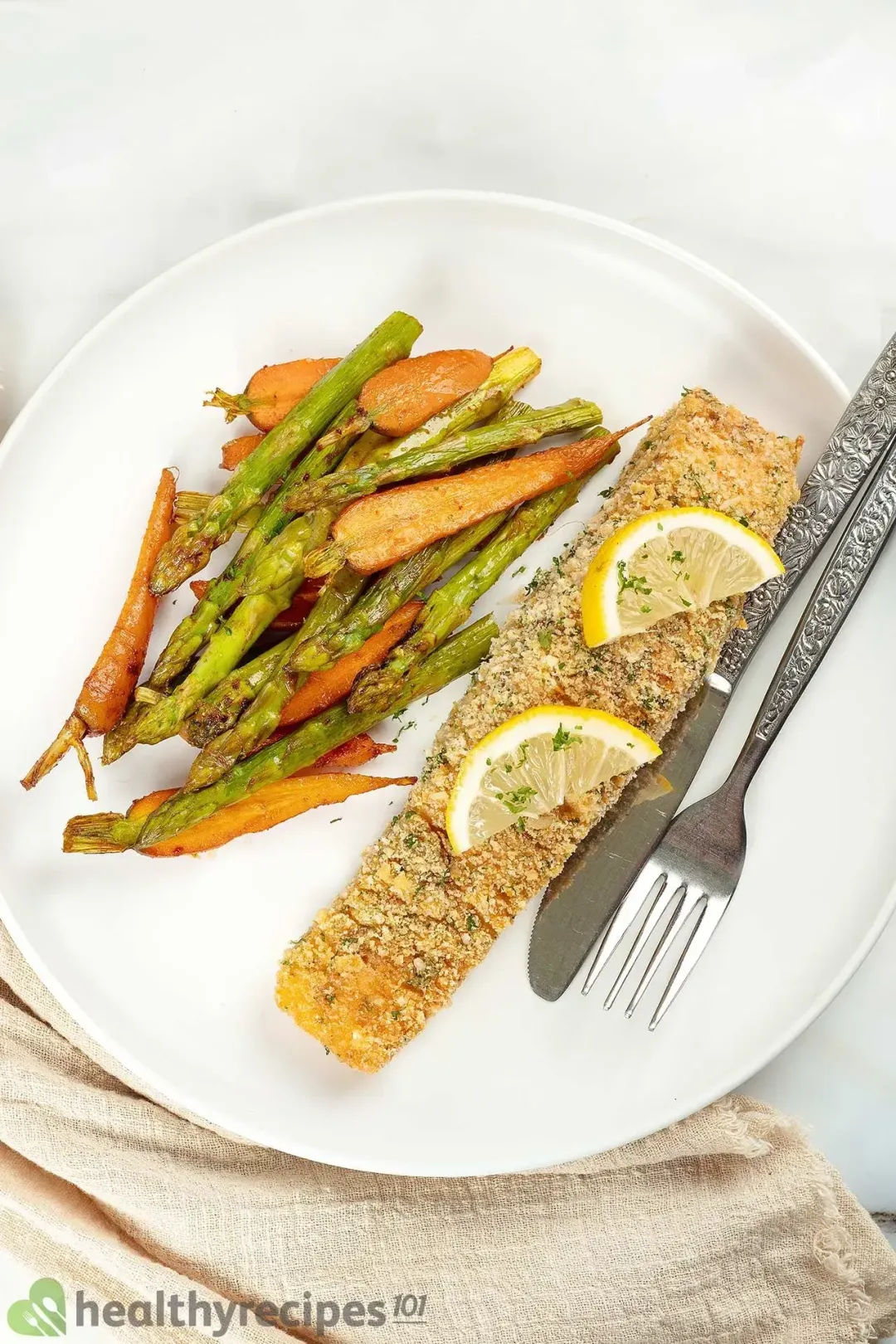 A white plate containing a salmon fillet crusted with breadcrumbs, some baby carrots, asparagus stalks, two lemon pieces, a dinner knife, and a fork