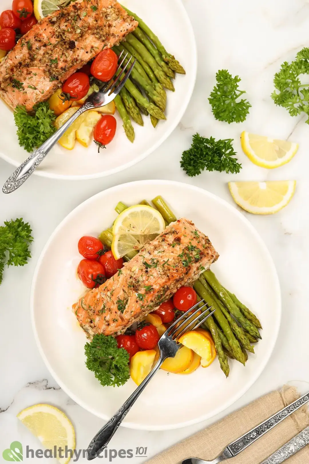 Two plates of baked salmon in foil with silver dining utensils surrounded by some fresh parsley and slices of lemon