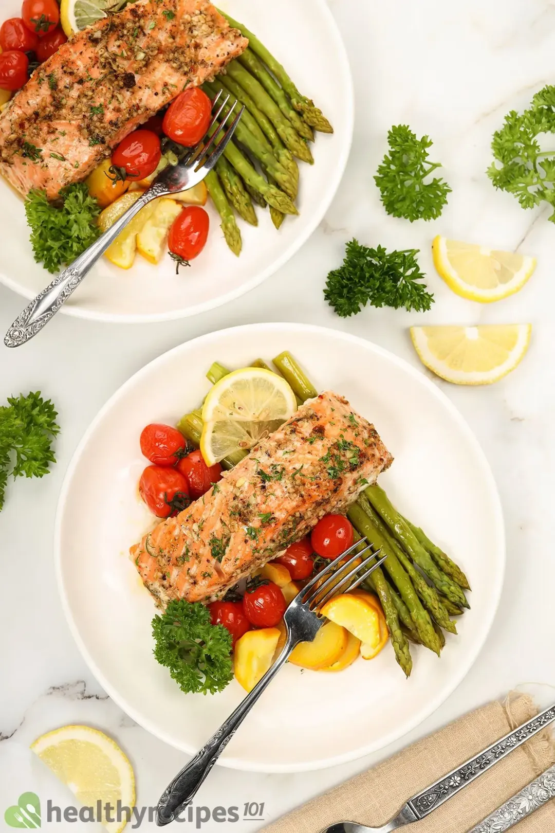 Two plates of baked salmon in foil with silver dining utensils surrounded by some fresh parsley and slices of lemon