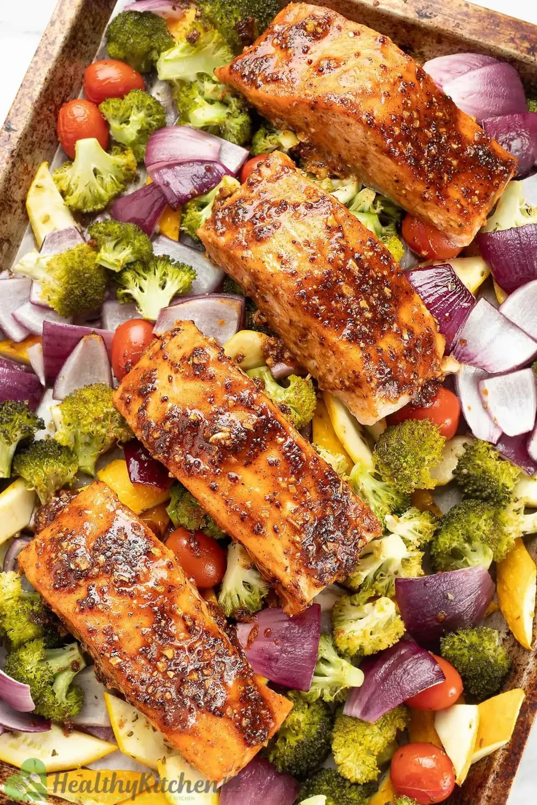A sheer pan containing colorful broccoli florets, red onion pieces, yellow zucchini pieces, cherry tomatoes, and salmon fillets covered in a glossy brown sauce