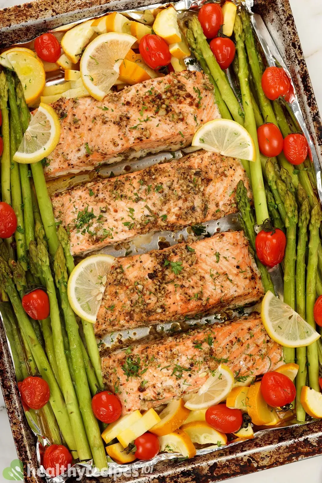 A baking tray lined with parchment paper full of seasoned salmon fillets, asparagus, cherry tomatoes, zucchini, and lemon slices