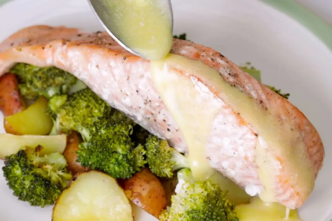 A spoon of buttery sauce is being poured on top of a baking salmon filet placed on top of some potato and broccoli on a white plate.