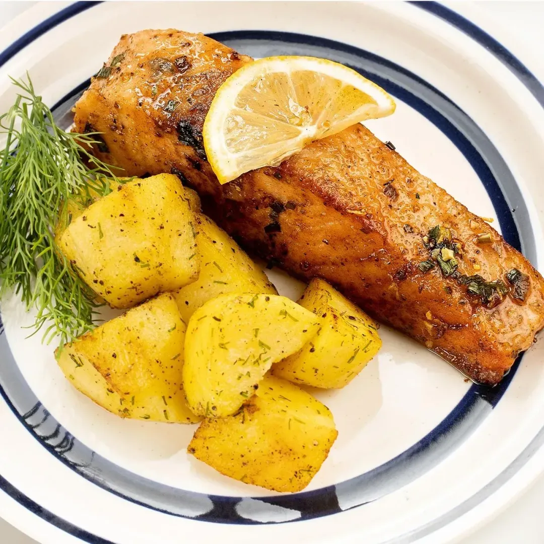 A white Chinese plate with a golden salmon filet topped with a lemon slice, potato wedges tossed with dill, and a dill sprig