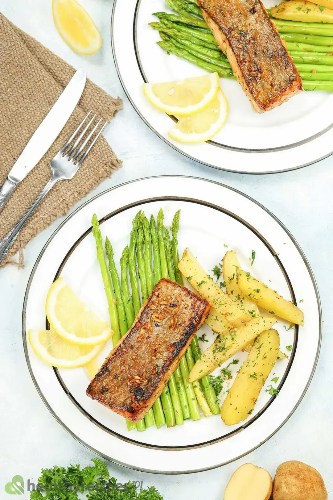A flatlay of a dinner knife, a fork, and two plates containing a cooked salmon fillet, potato wedges, asparagus stalks, and lemon slices
