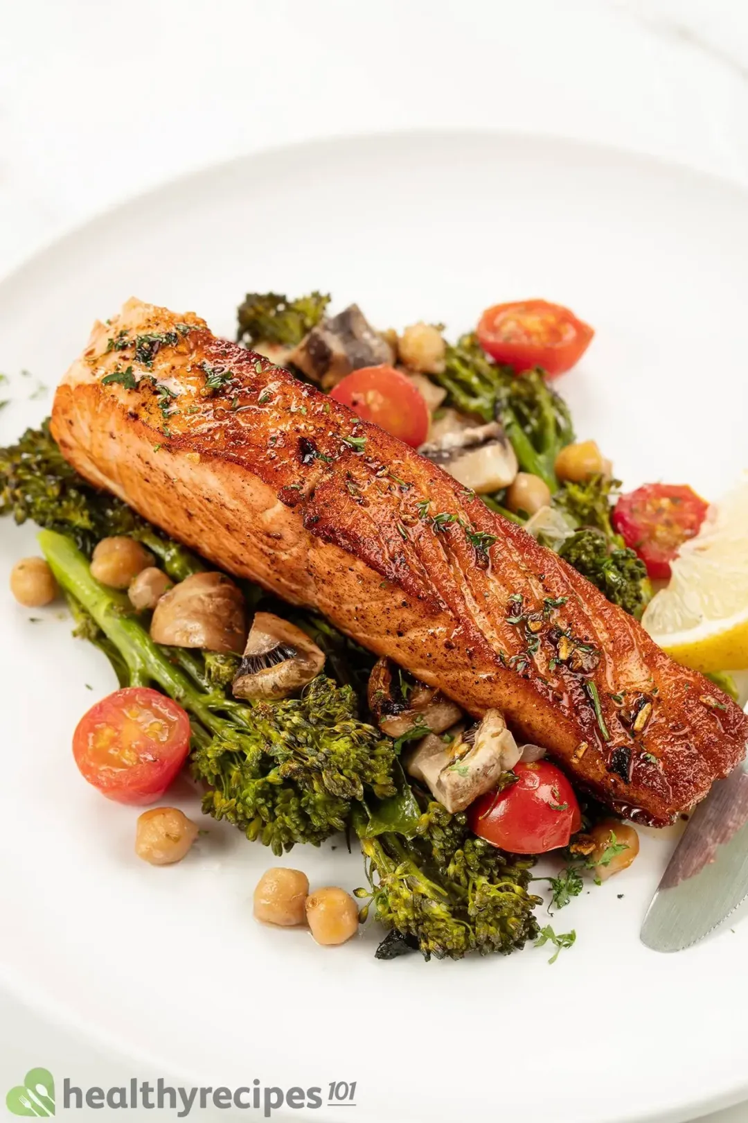 A white plate containing a pan-seared salmon fillet laid on a bed of broccolini, halved cherry tomatoes, cooked mushrooms, and chickpeas