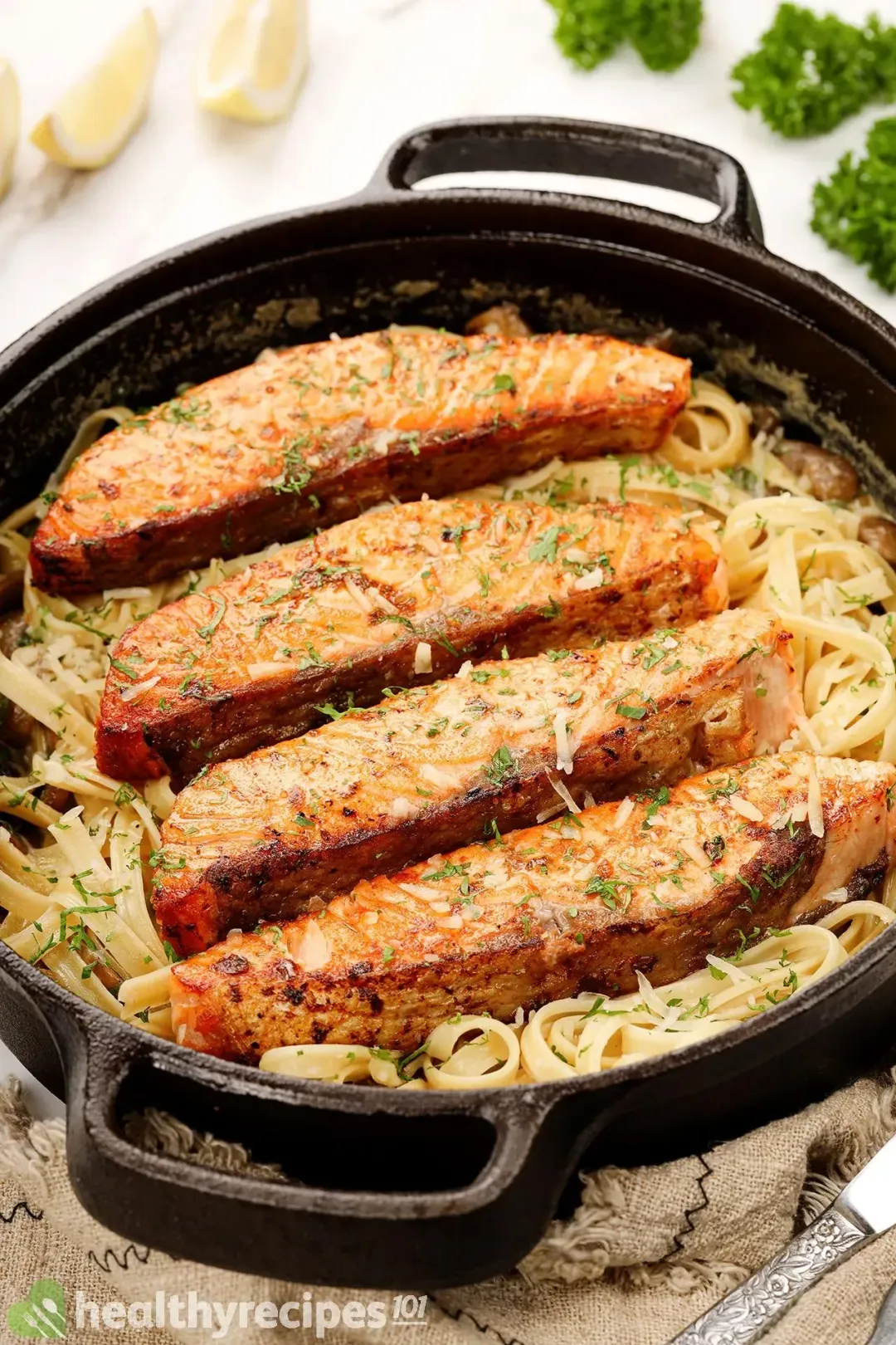 A skillet containing four bright orange cooked salmon fillets sprinkled with chopped parsley laid on a bed of fettuccine