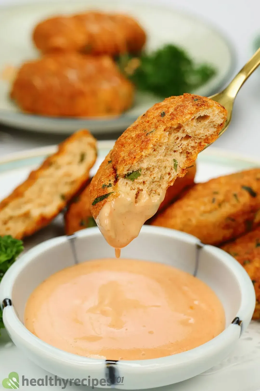 A close up picture of a piece of air fryer salmon patty dipped in a orange-colored sauce in the background of a plate with other patties.