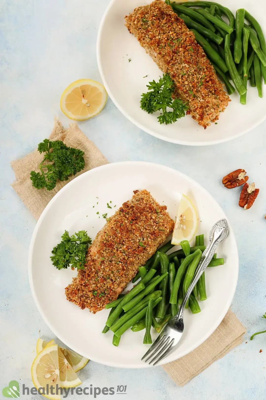 Two dishes represent two complete servings, on each there are one filet of pecan-crusted salmon, cooked green beans, parsley, lemon slices, and a food-use serving fork.