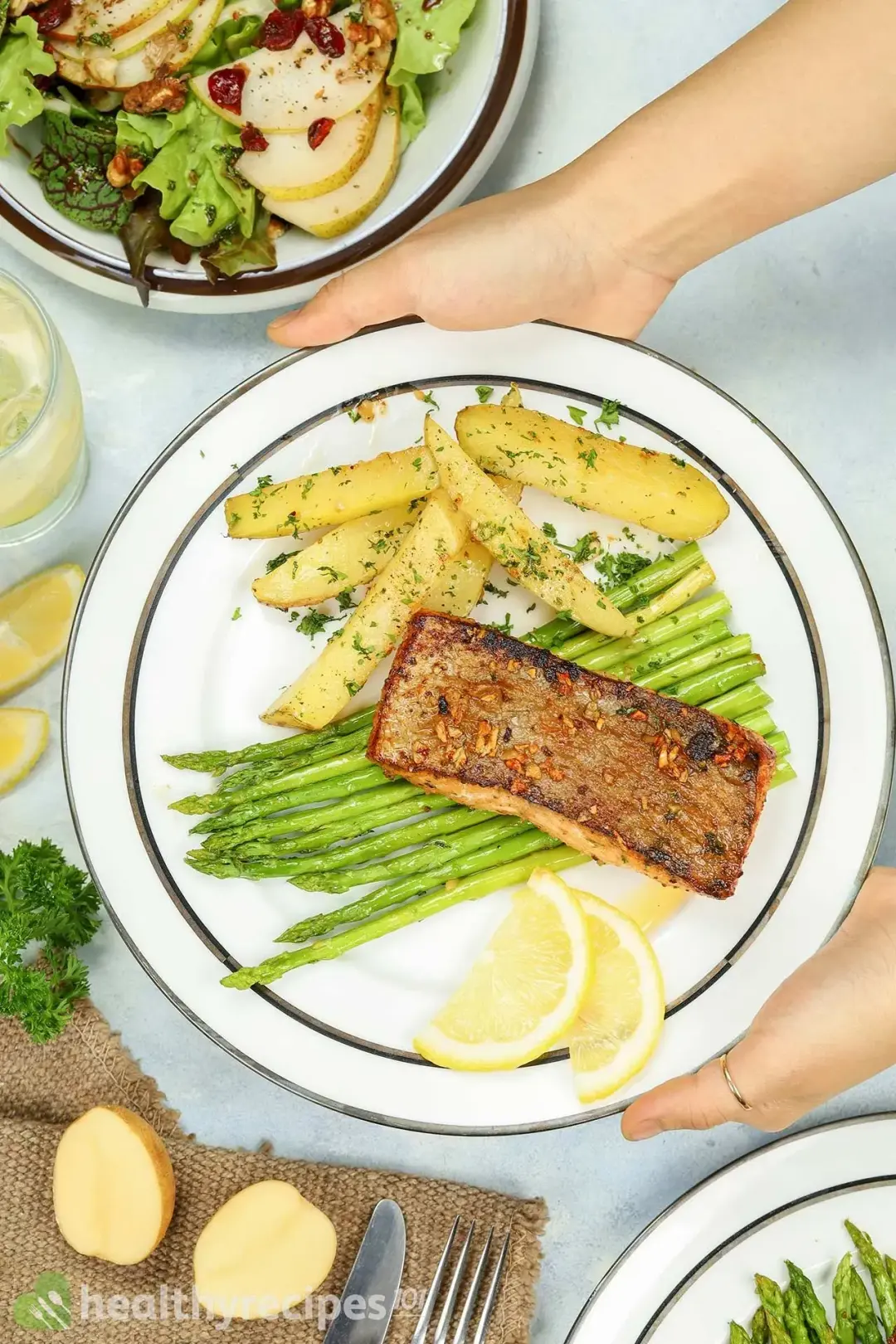 Two hands holding a plate containing a piece of pan-seared salmon, asparagus, potato wedges, and lemon slices