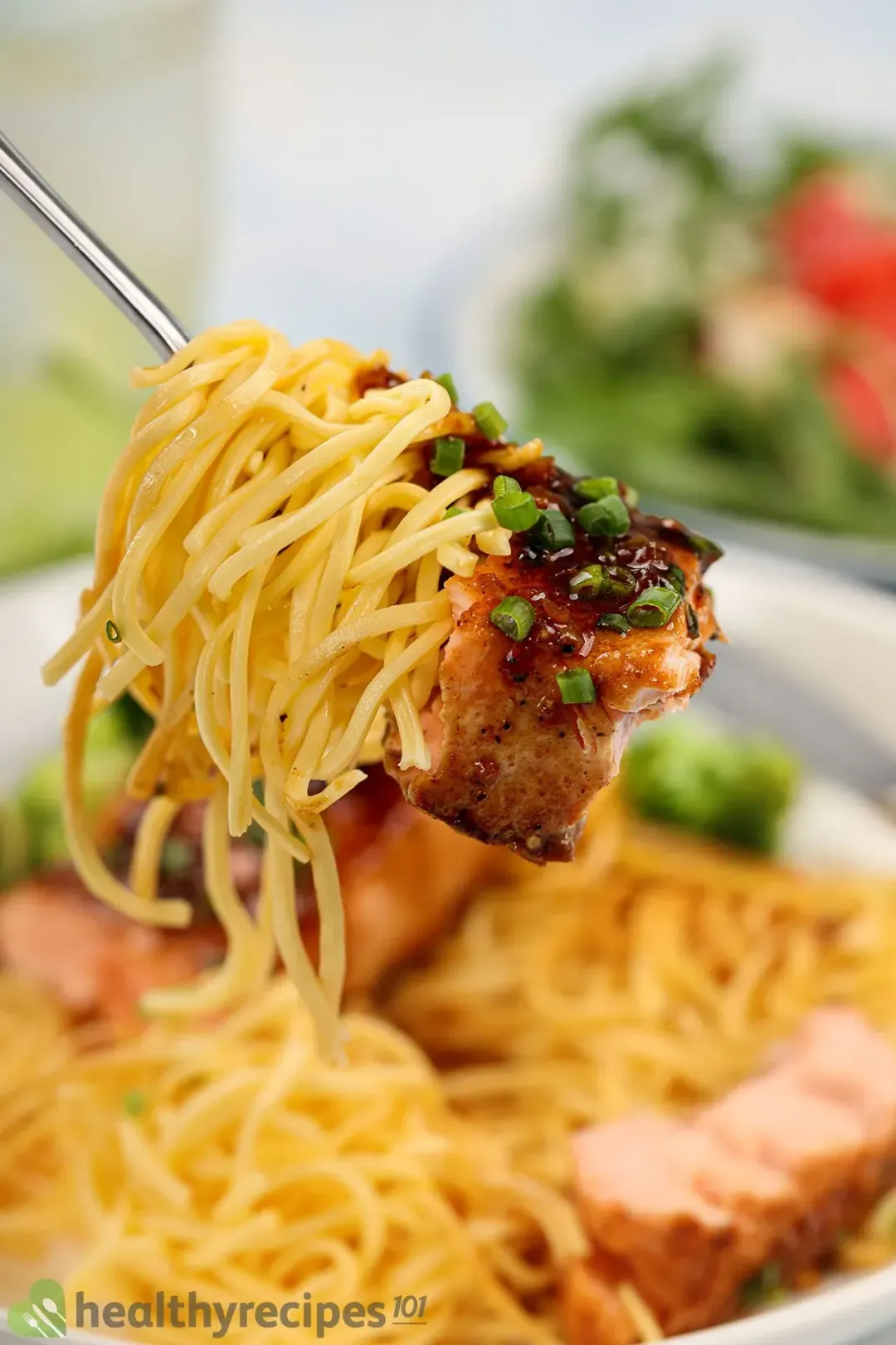 A forkful of cooked egg noodles and honey garlic salmon with a plate of honey garlic salmon and watermelon arugula salad on the background