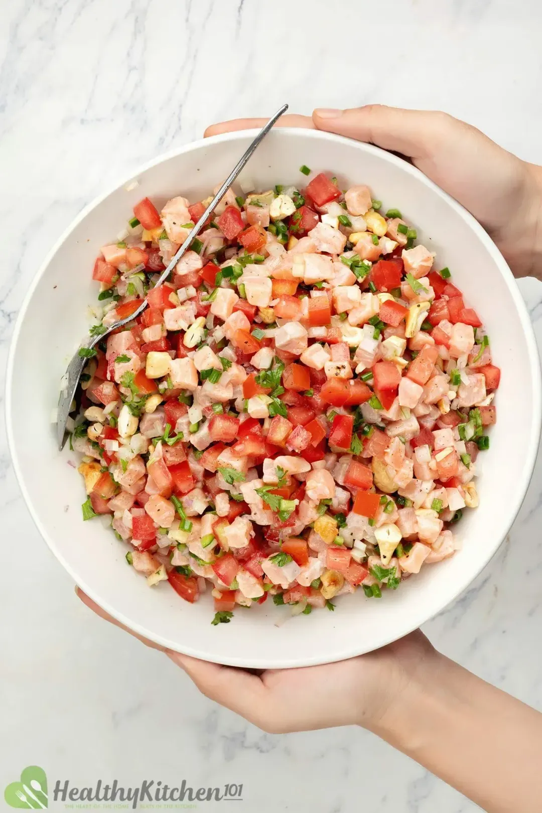 Two hands holding a large plate of Lomi Lomi salmon salad with salmon cubes, chopped tomatoes, and chopped herbs